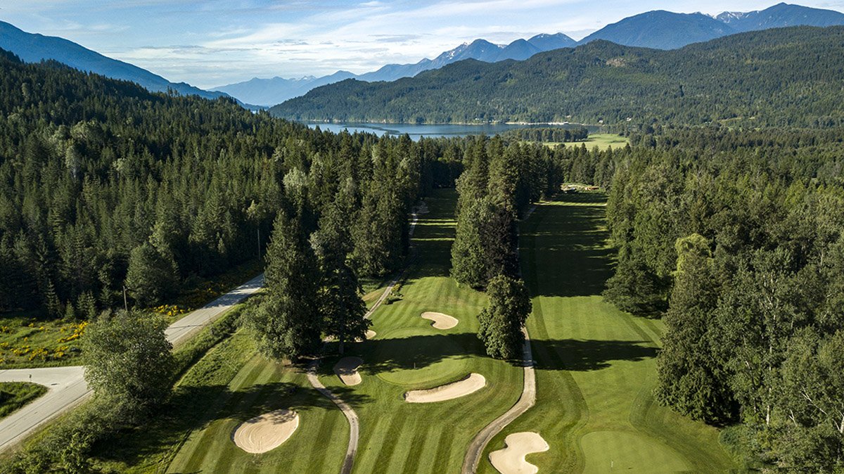 A scenic view of a golf course nestled in a lush, green forest with mountains and a body of water in the background, under a partly cloudy sky.