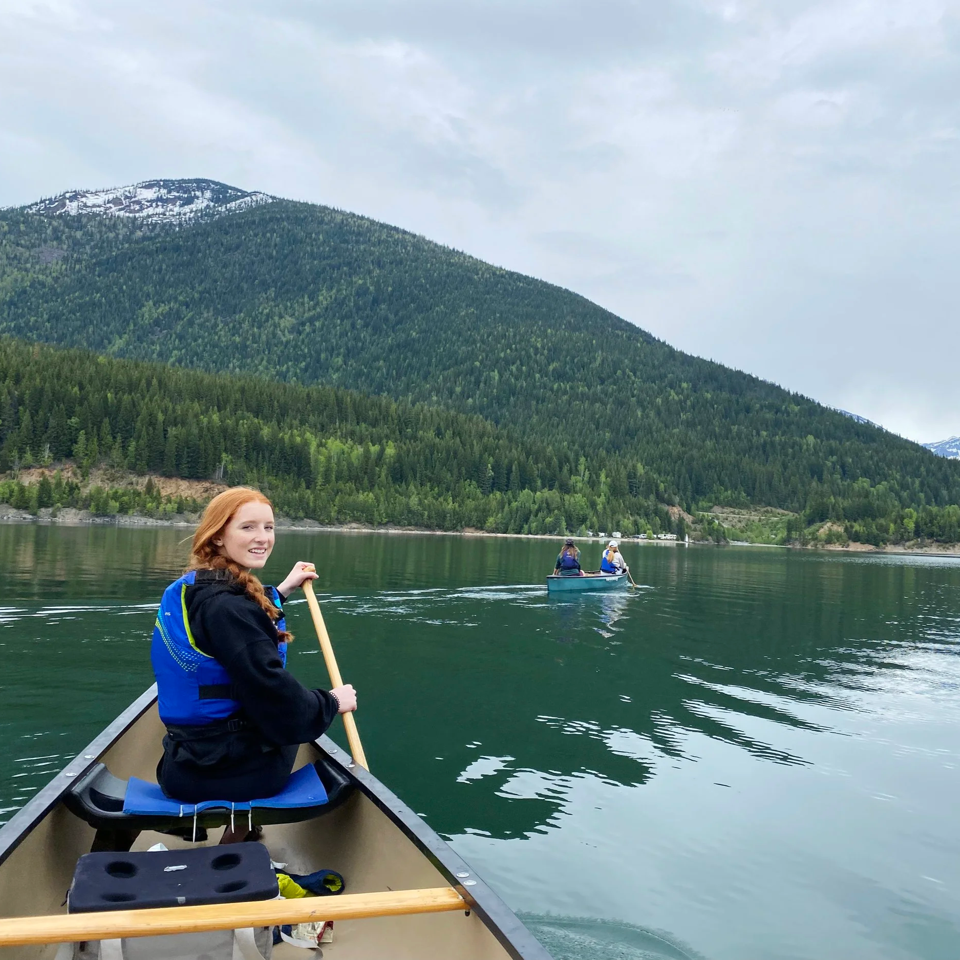 A young woman with red hair smiling and sitting in a canoe on a calm lake, wearing a black jacket and a blue life vest. She is holding a paddle, and there are other people in a canoe in the background. The lake is surrounded by lush green trees and a mountainous landscape with patches of snow under a cloudy sky.