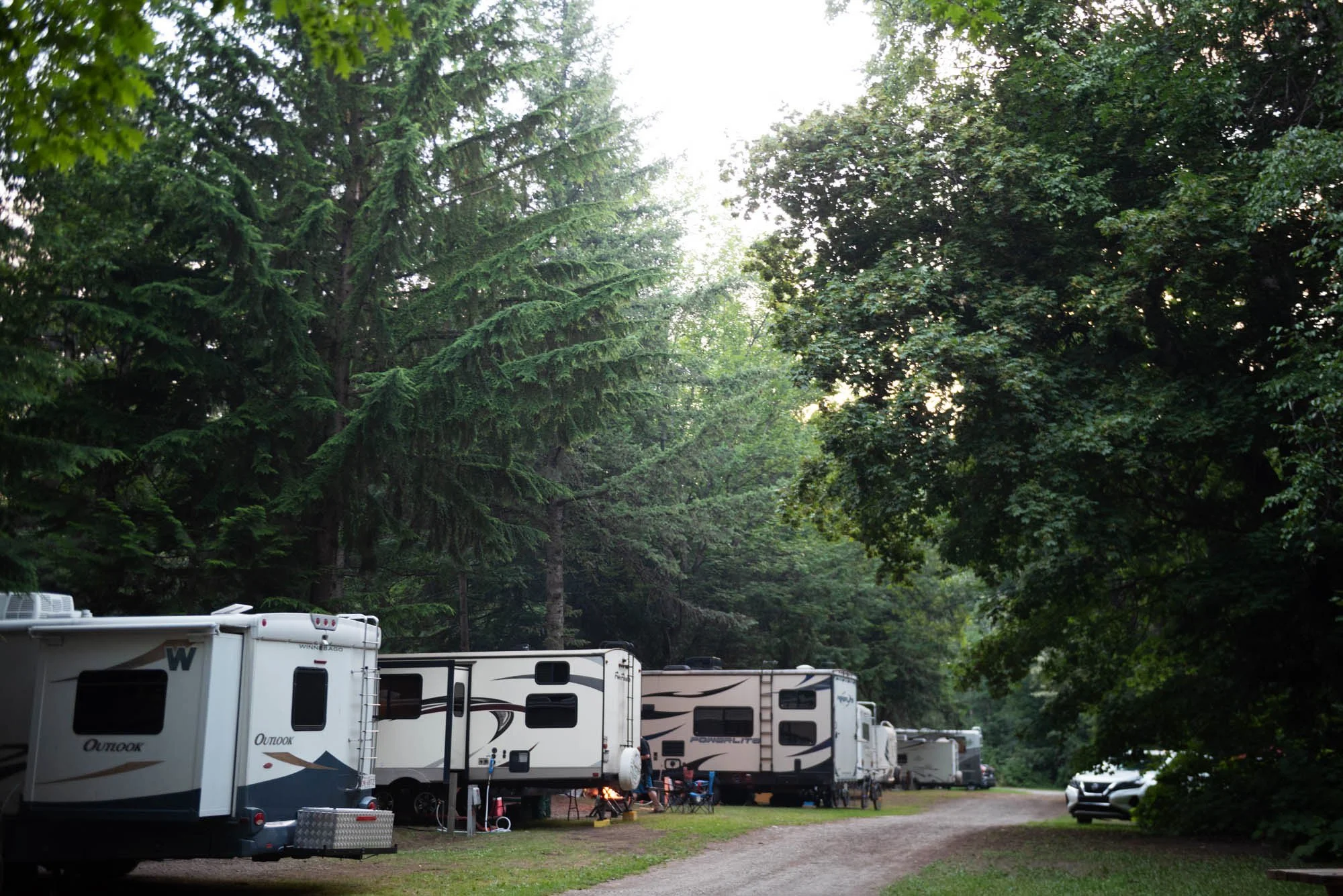 A parking area for RVs and trailers in a wooded area with tall green trees, some sunlight filtering through the foliage, and a few cars parked on the dirt ground.