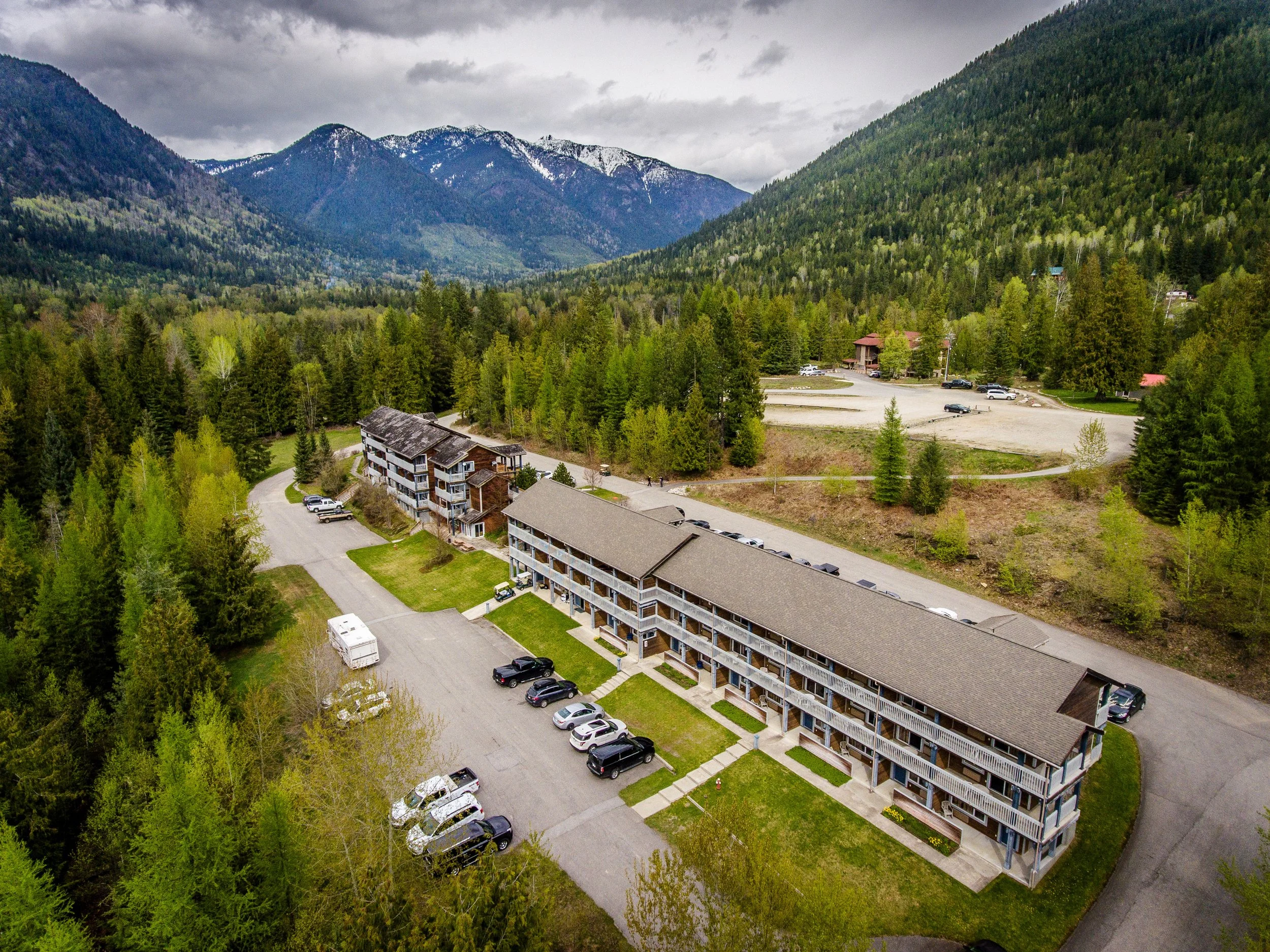 Aerial view of a mountain landscape with green trees, snow-capped peaks, and a small residential area with parked cars.
