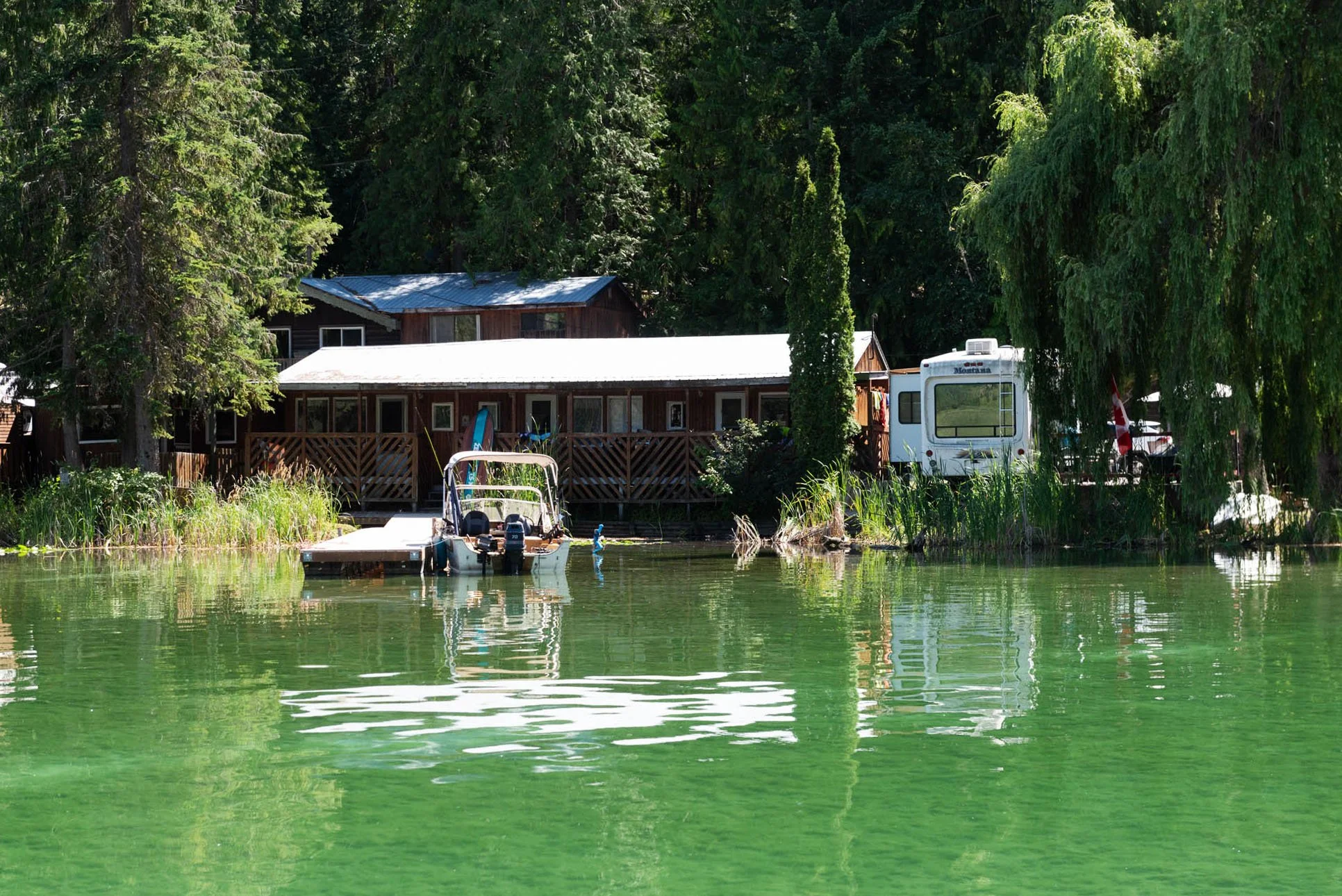 A lakeside scene with a wooden house and a camper surrounded by green trees. A boat with outboard motors is docked at a small pier in the water. The water is slightly rippled, reflecting the greenery and structures.