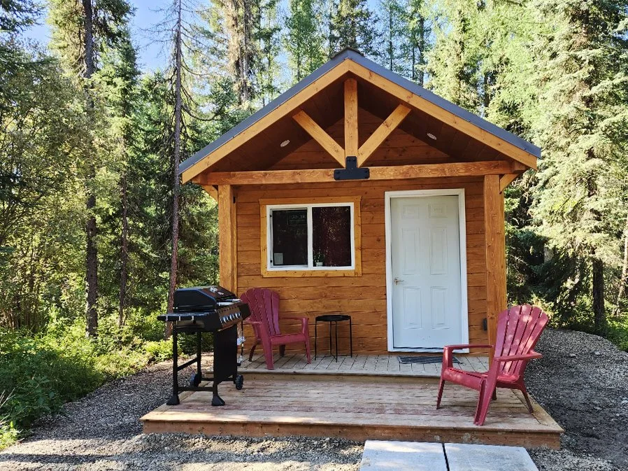 Small wooden cabin with a porch, two red Adirondack chairs, a black gas grill, and a small black side table, surrounded by green trees.