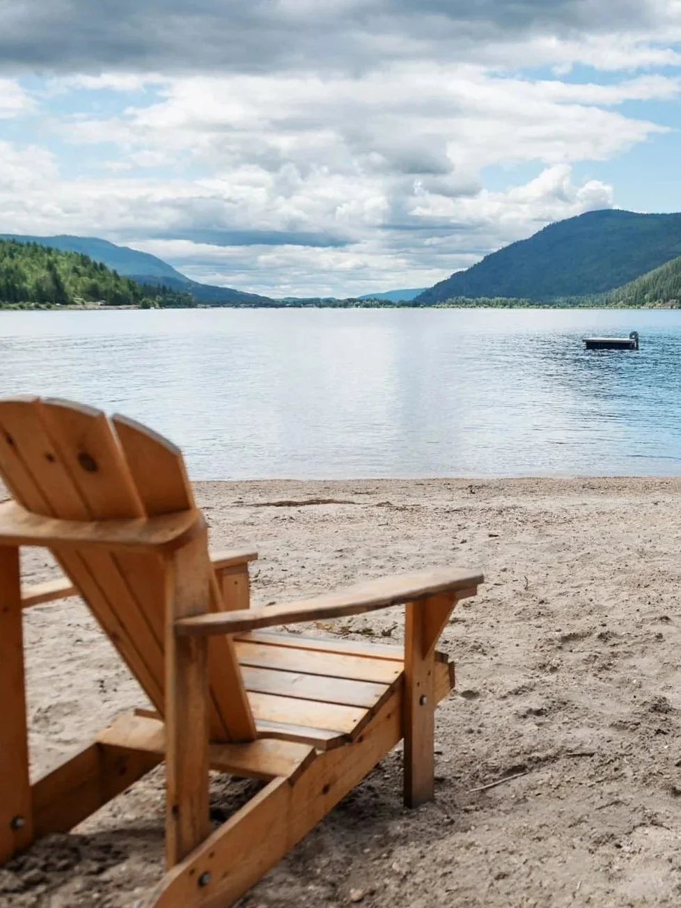 A wooden Adirondack chair on a sandy beach overlooking a calm lake with hills and mountains in the distance under a partly cloudy sky.