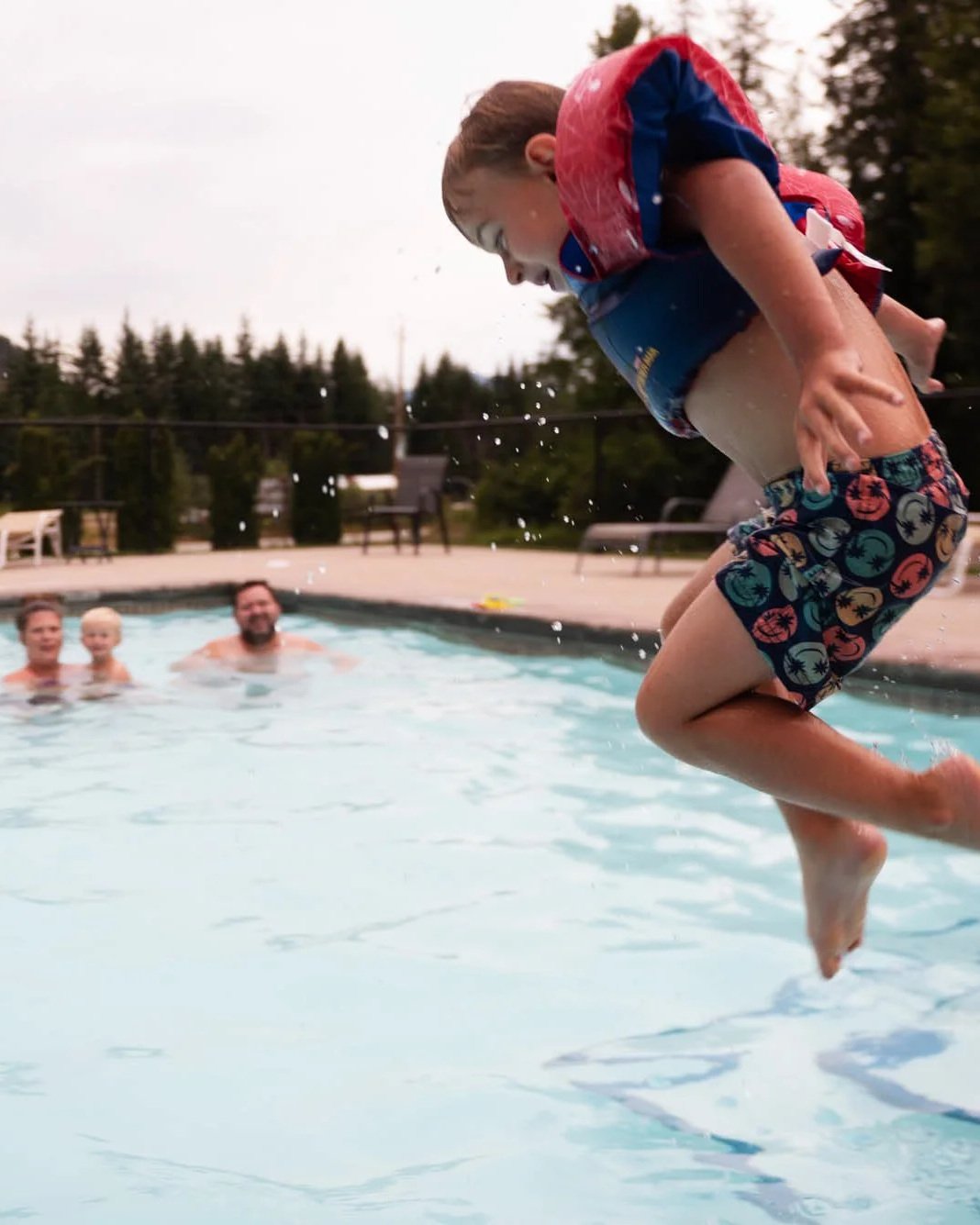 Child jumping into a swimming pool with family in the background, outdoors, on a cloudy day.