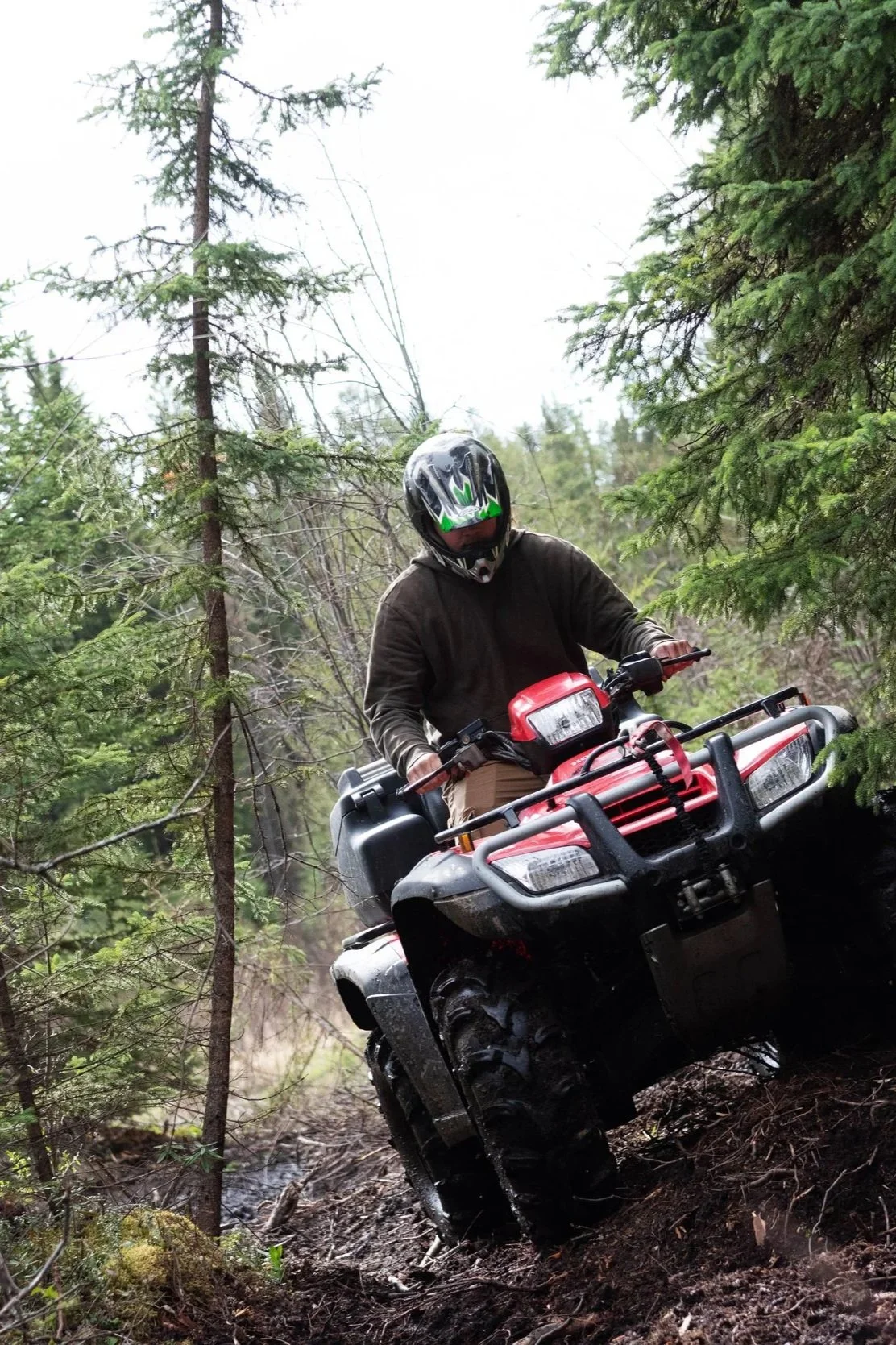 Person riding a red all-terrain vehicle (ATV) through a dense forest, wearing a helmet and brown jacket.