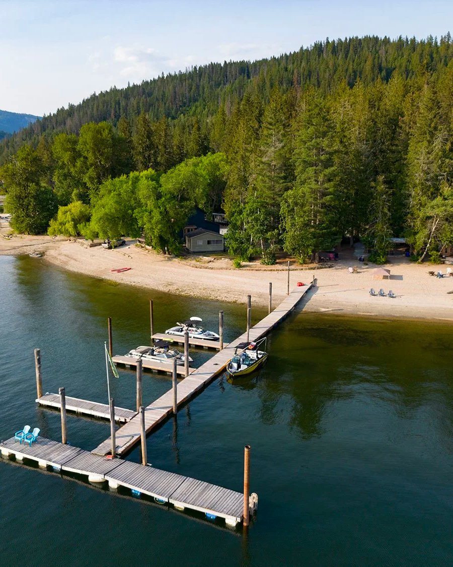 A dock extending into a calm body of water, with boats tied to the dock, and a beach with trees and summer chairs in the background.