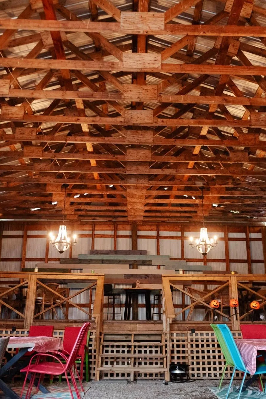 Interior of a rustic barn with wooden beams, chandelier lighting, and a small stage or platform at the back. There are colorful chairs and tables in the foreground, and jack-o'-lantern decorations on the railing.