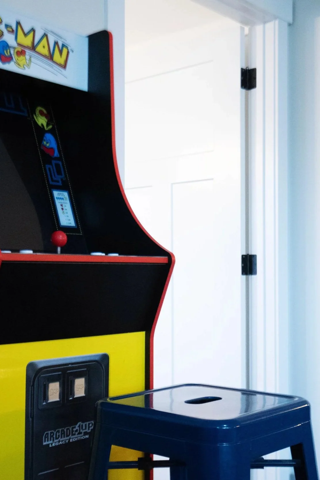 An arcade game machine, part of the Pac-Man arcade game, with a black screen, joystick, and buttons, positioned near a white door and a small table or stand in a room.