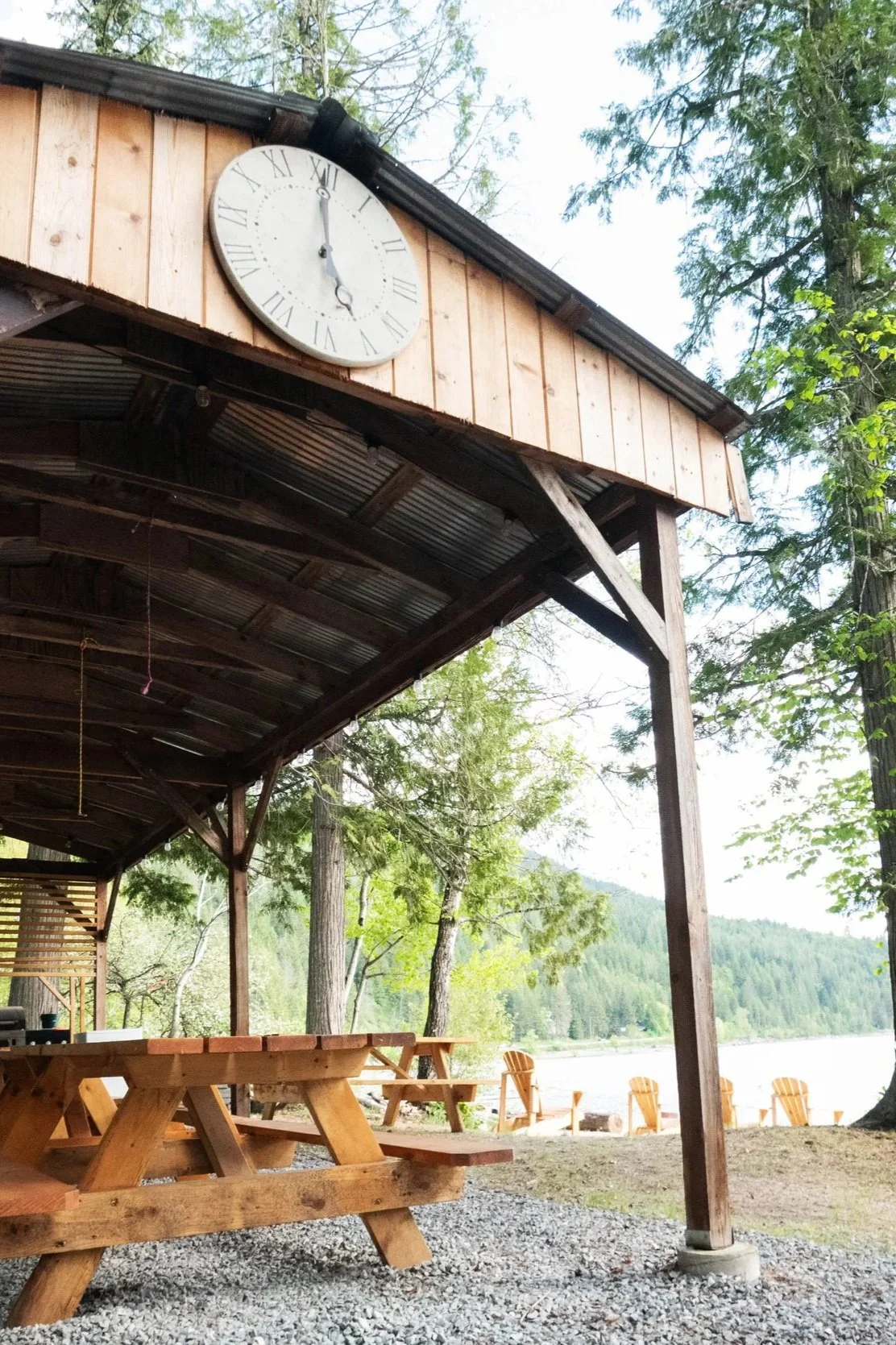 Wooden outdoor shelter with a round clock showing 12:30, picnic tables, Adirondack chairs, trees, and a lake in the background.