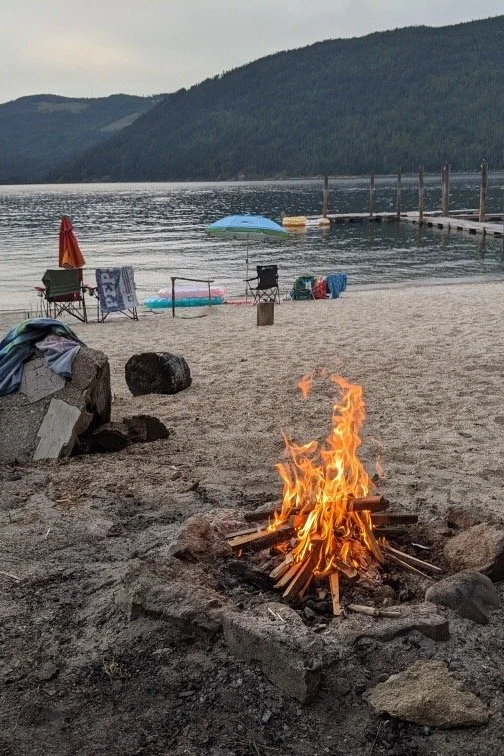 A campfire burning on a sandy beach near a lake with a dock, chairs, umbrellas, and mountains in the background.