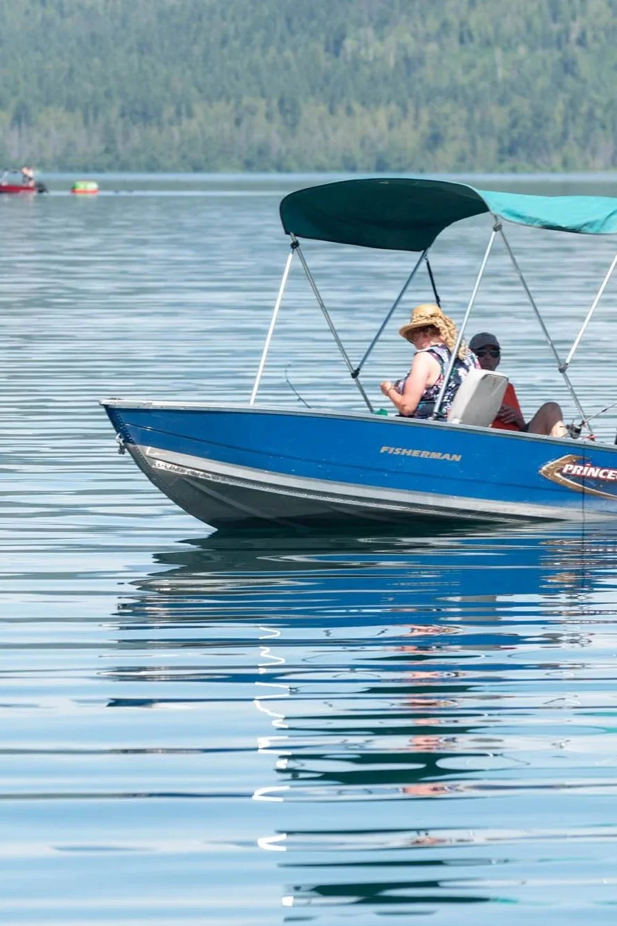 A blue boat named Fisherman with a canopy, carrying two people, one wearing a straw hat, on a calm lake with a forested shoreline in the background.