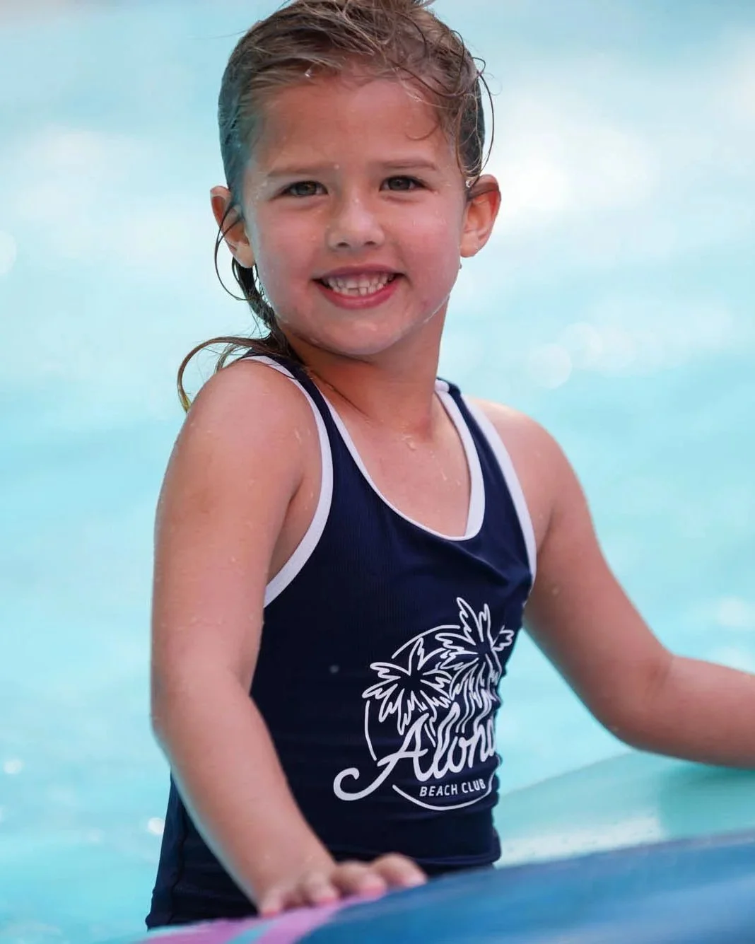 Young girl smiling in a swimming pool, wearing a navy blue swimsuit with white trim and a palm tree logo that says Alexis Beach Club.