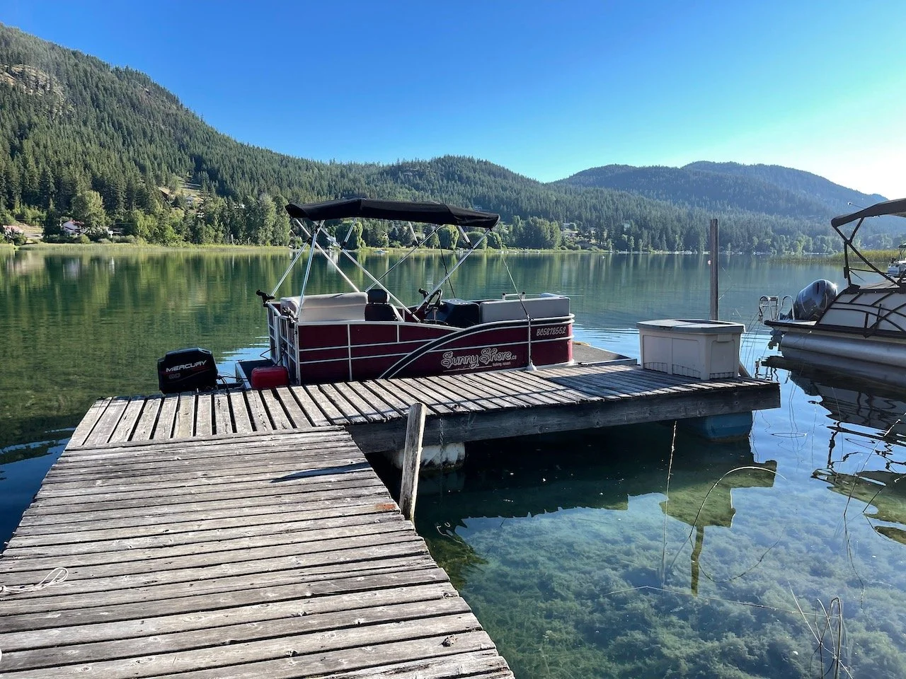 A wooden dock extending into a calm lake with a red pontoon boat and another boat in the background, surrounded by green mountains under a clear blue sky.