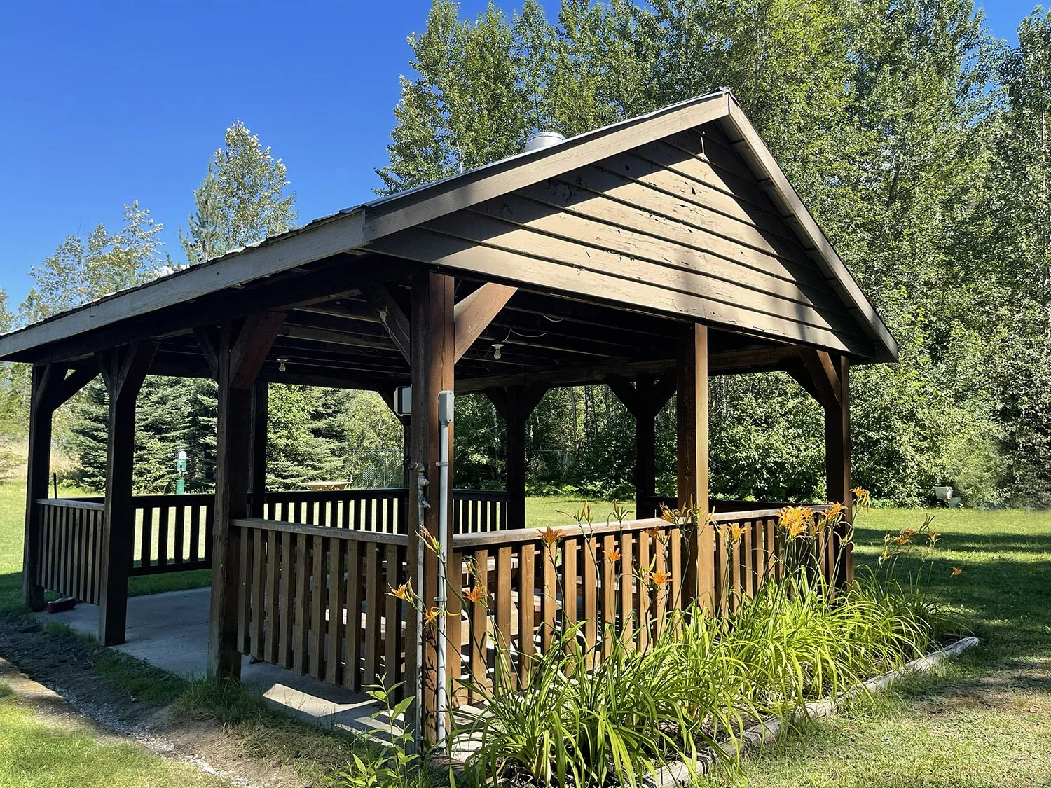A wooden gazebo with a slanted roof, open sides, and a railing, situated in a grassy outdoor area with trees and blue sky in the background.