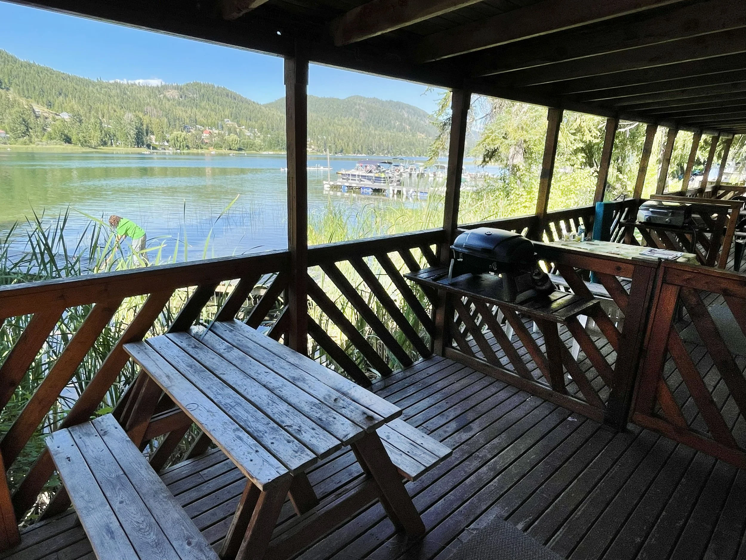 View from a covered wooden porch overlooking a lake, with docked boats, surrounding green hills, and a person in a green shirt near the water.