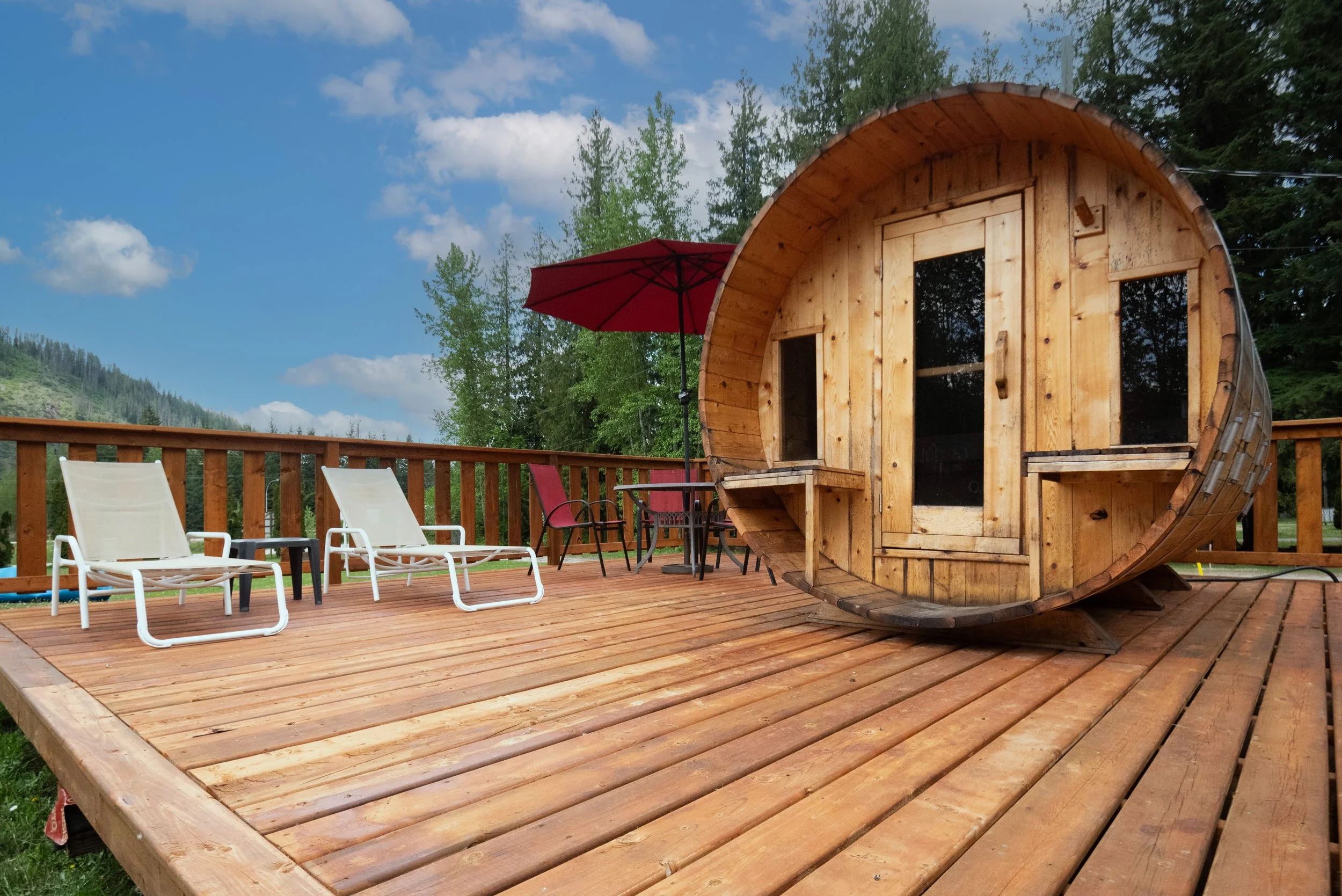 Wooden barrel-shaped tiny house on a deck with outdoor furniture, a red umbrella, and scenic trees and mountains in the background.