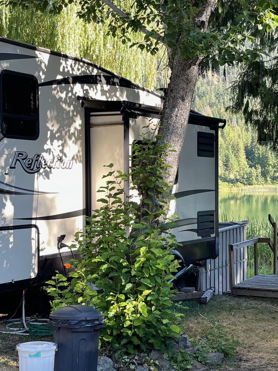 Travel trailer parked near a lake with trees and greenery, a small wooden deck, and trash bins in the foreground.