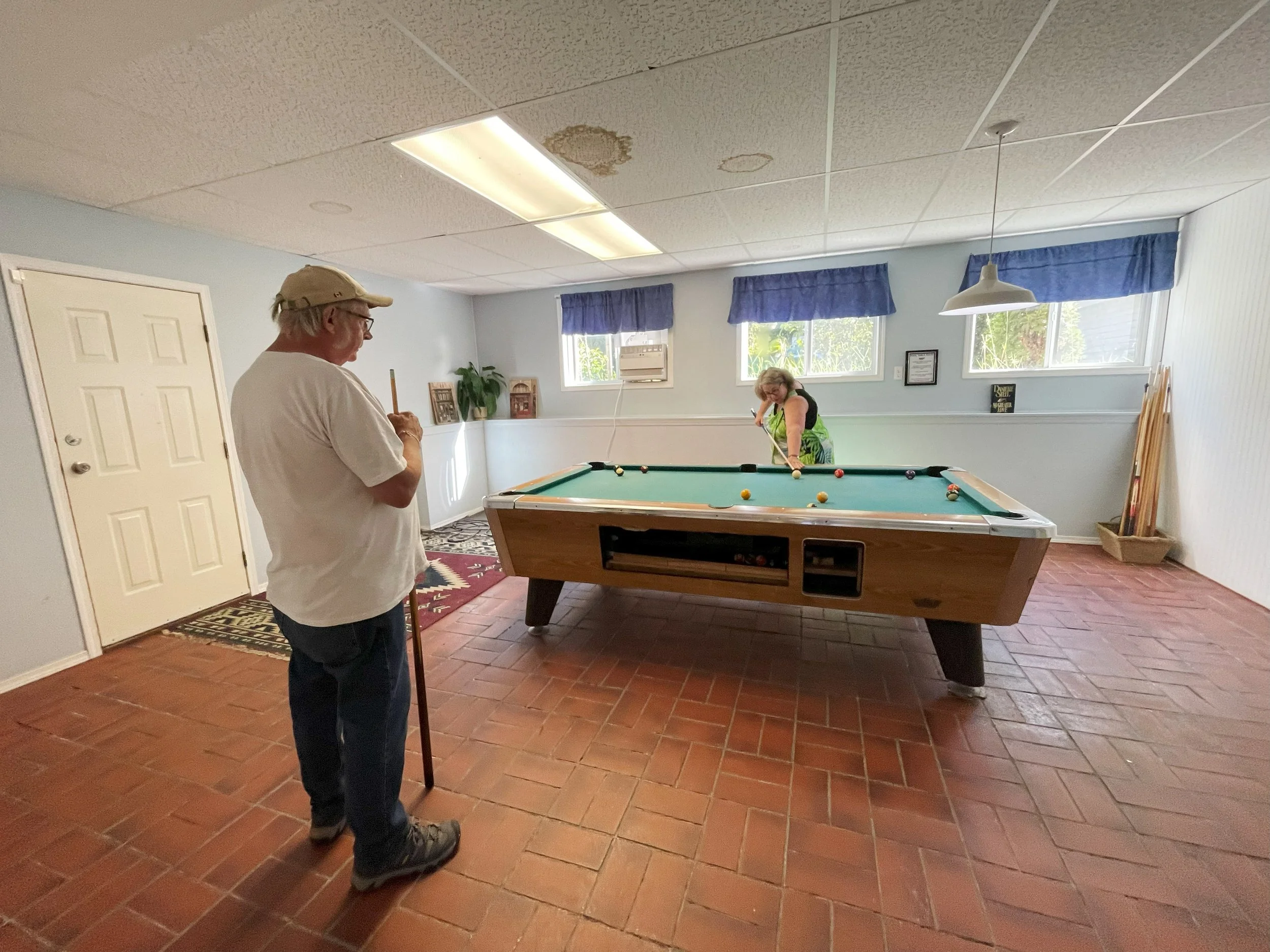 An elderly man in glasses and a beige cap standing by a pool table, while a woman in glasses and a green apron is preparing to take a shot in a room with light blue walls, two windows with blue curtains, and a tiled floor.