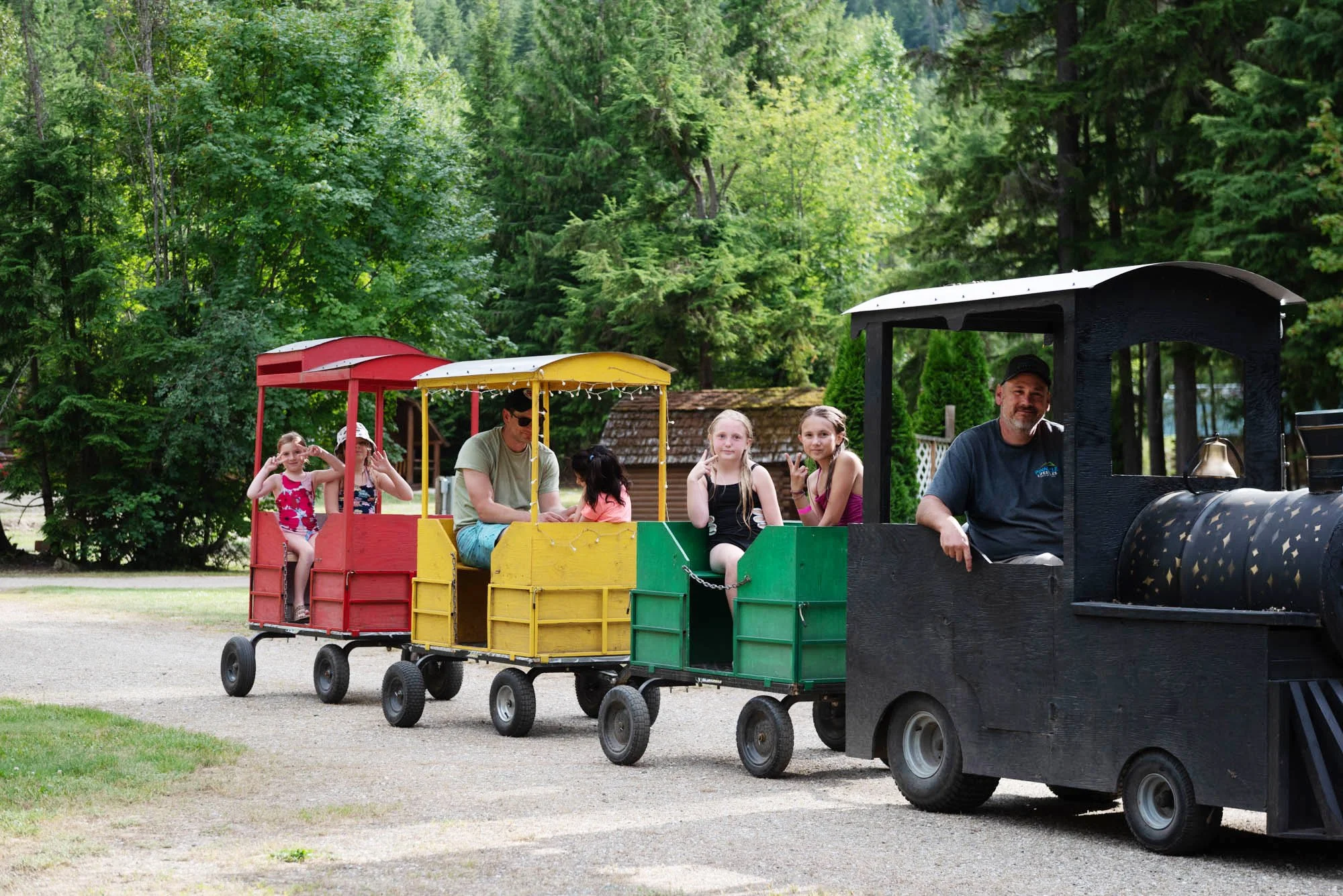 People riding a colorful train ride in a park surrounded by tall green trees.