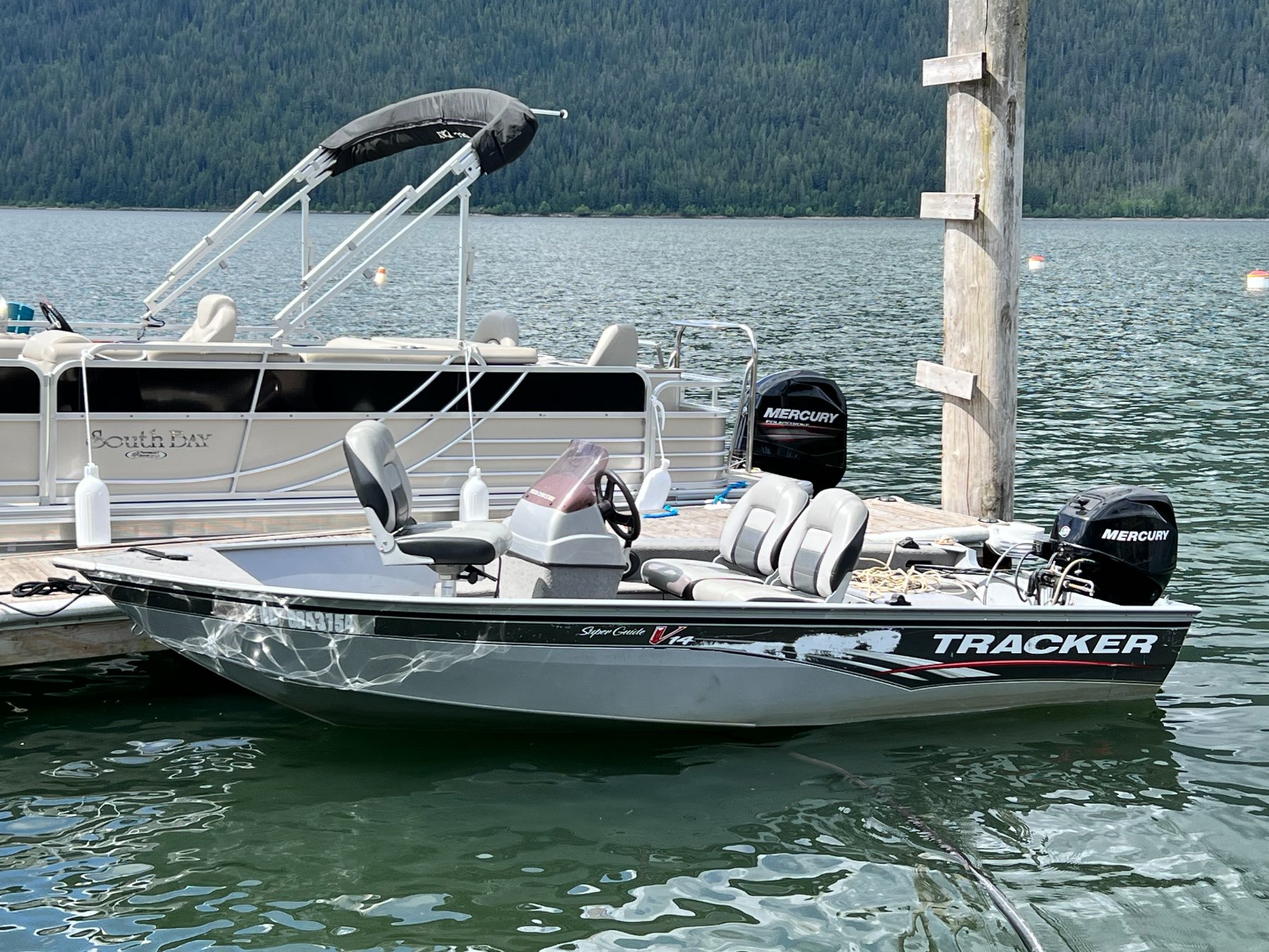 Two boats docked at a pier on a lake with forested mountains in the background. The boats have outboard motors and are equipped with seats and canopies.