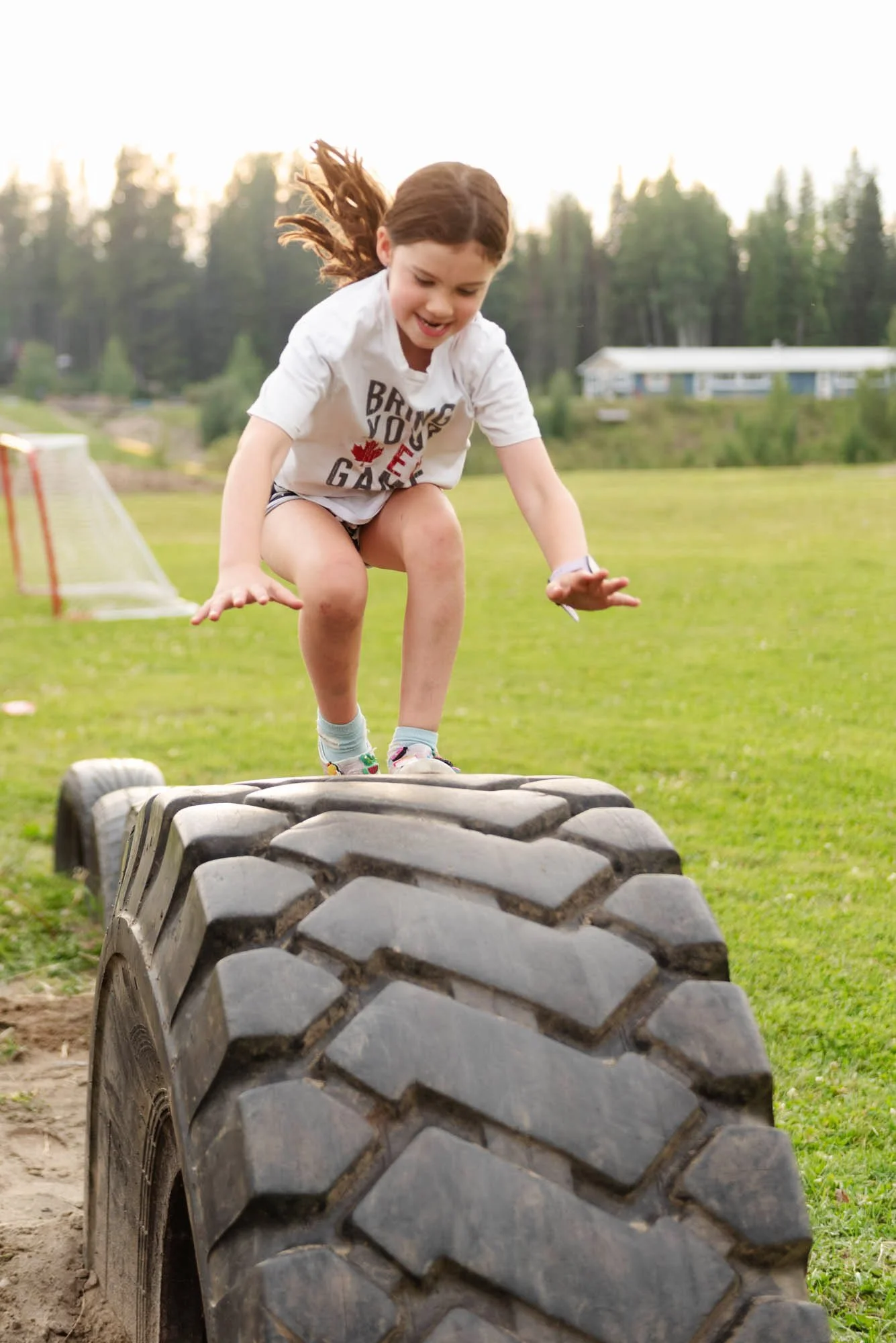 A young girl with brown hair, wearing a white T-shirt with text and shorts, jumping over a large tire on a grassy field during daytime.