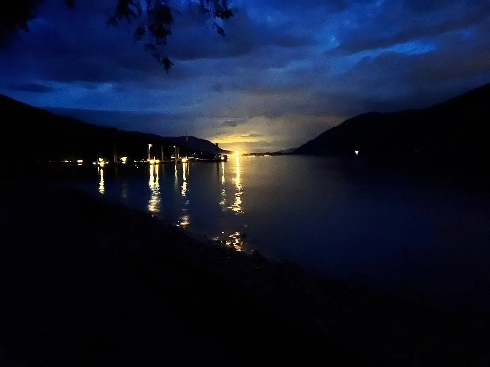 Nighttime landscape of a lake surrounded by hills, with dark clouds overhead and a bright light on the horizon reflecting on the water. Several boats are docked on the left side of the lake, with some lights shining from nearby buildings.
