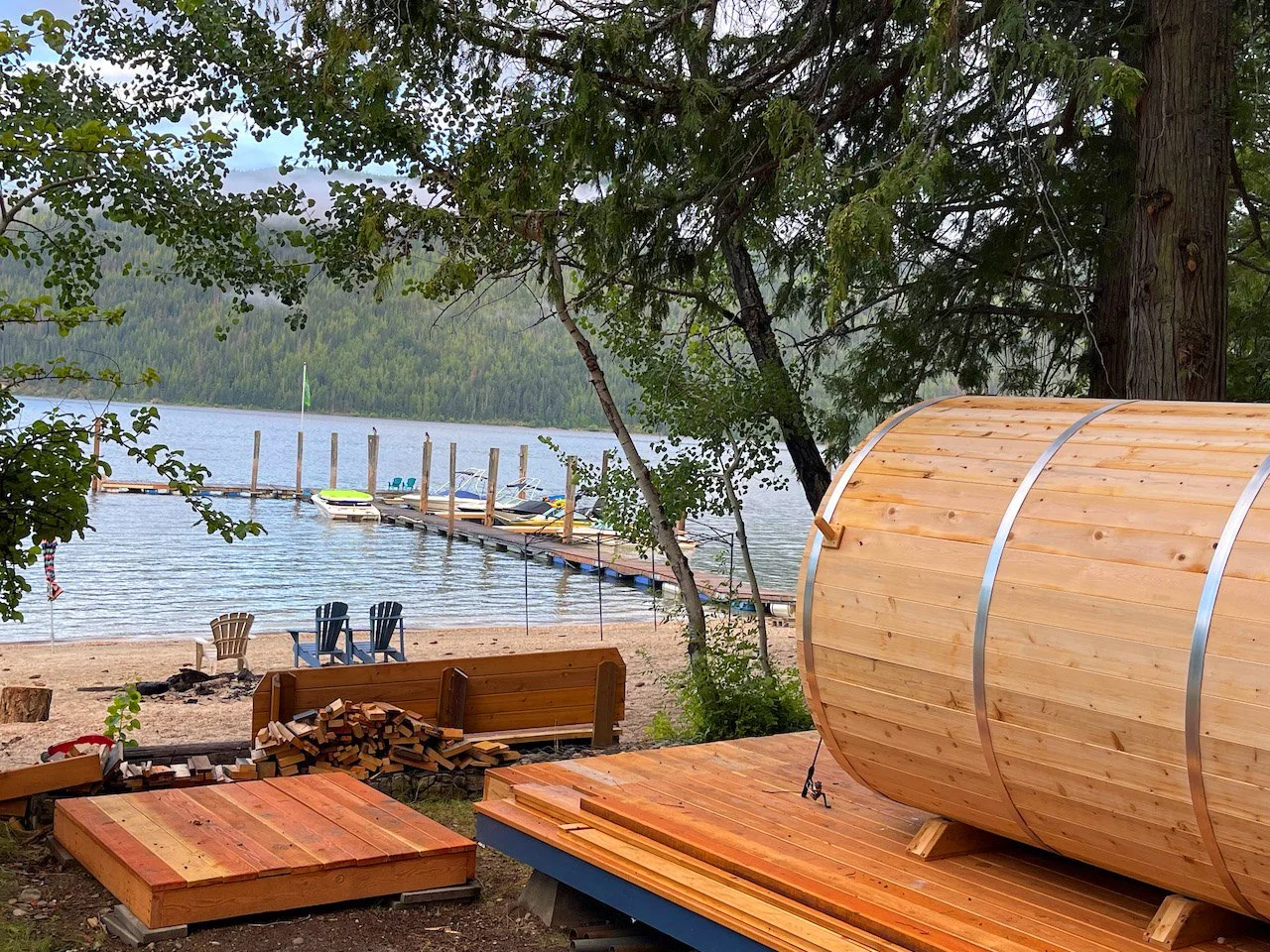 A lakeside scene showing a dock with boats, two Adirondack chairs, a firepit, stacked wood, and a large wooden barrel sauna under trees near the water.