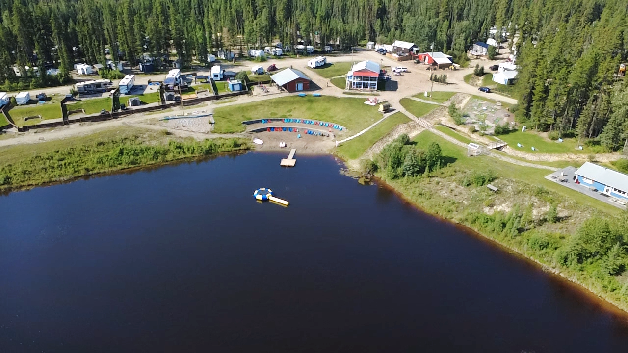 Aerial view of a lakeside campground with RVs, small buildings, and a floating water park on a dark lake, surrounded by trees at Pinnacle Trails Resort