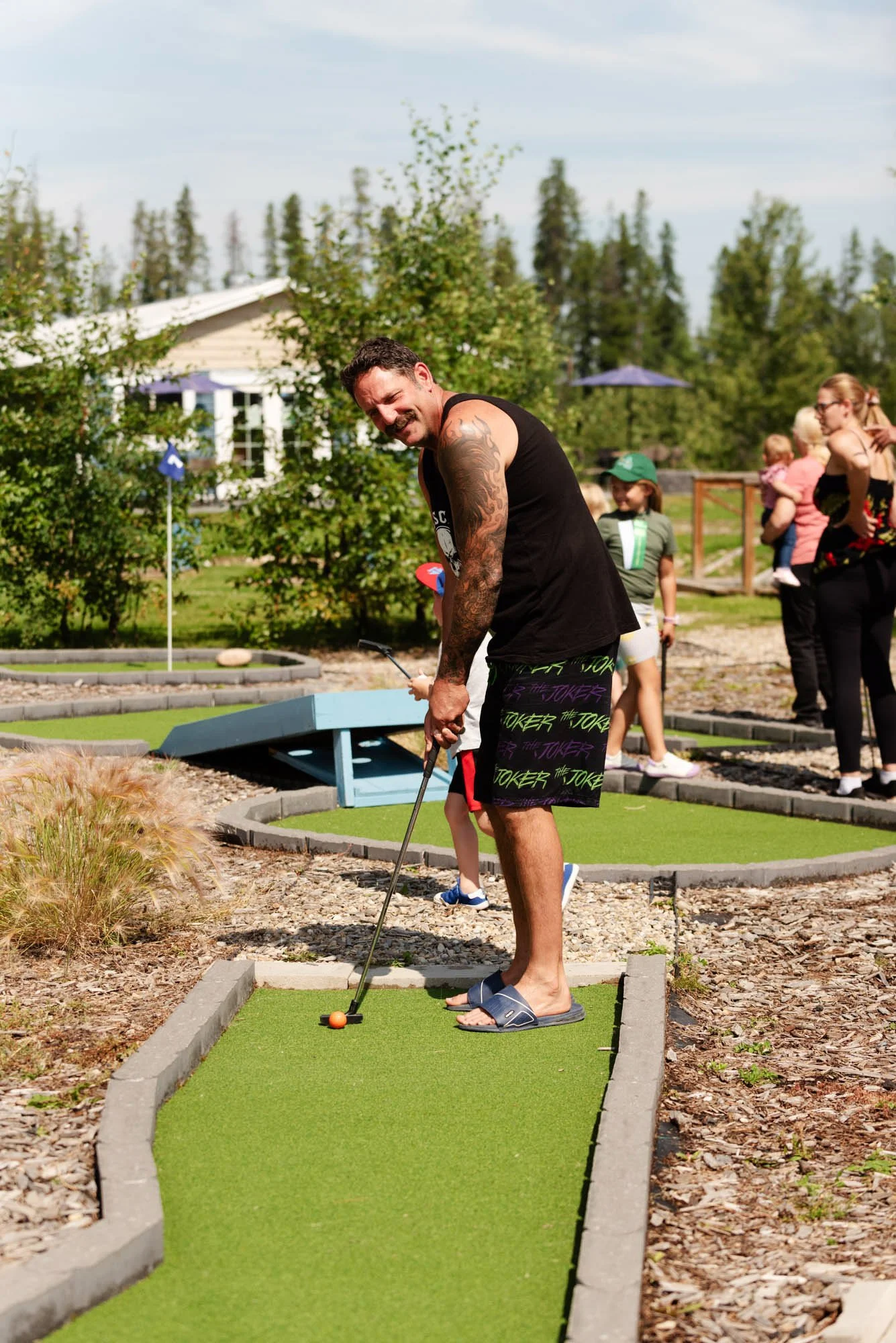 A man in a black sleeveless shirt and camo shorts is playing miniature golf. He is barefoot and wearing slippers. He is about to hit an orange golf ball with a putter on a small green course while smiling. In the background, there are other people, trees, and houses.