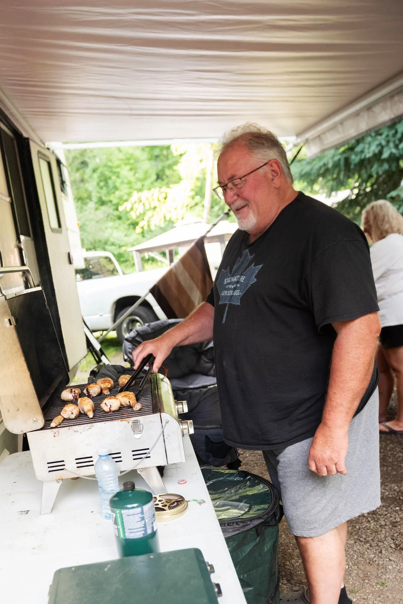 A man with gray hair, glasses, and a beard grilling hot dogs on a barbecue grill outdoors. He is wearing a black T-shirt with a maple leaf logo and gray shorts. There are people sitting in the background under trees.
