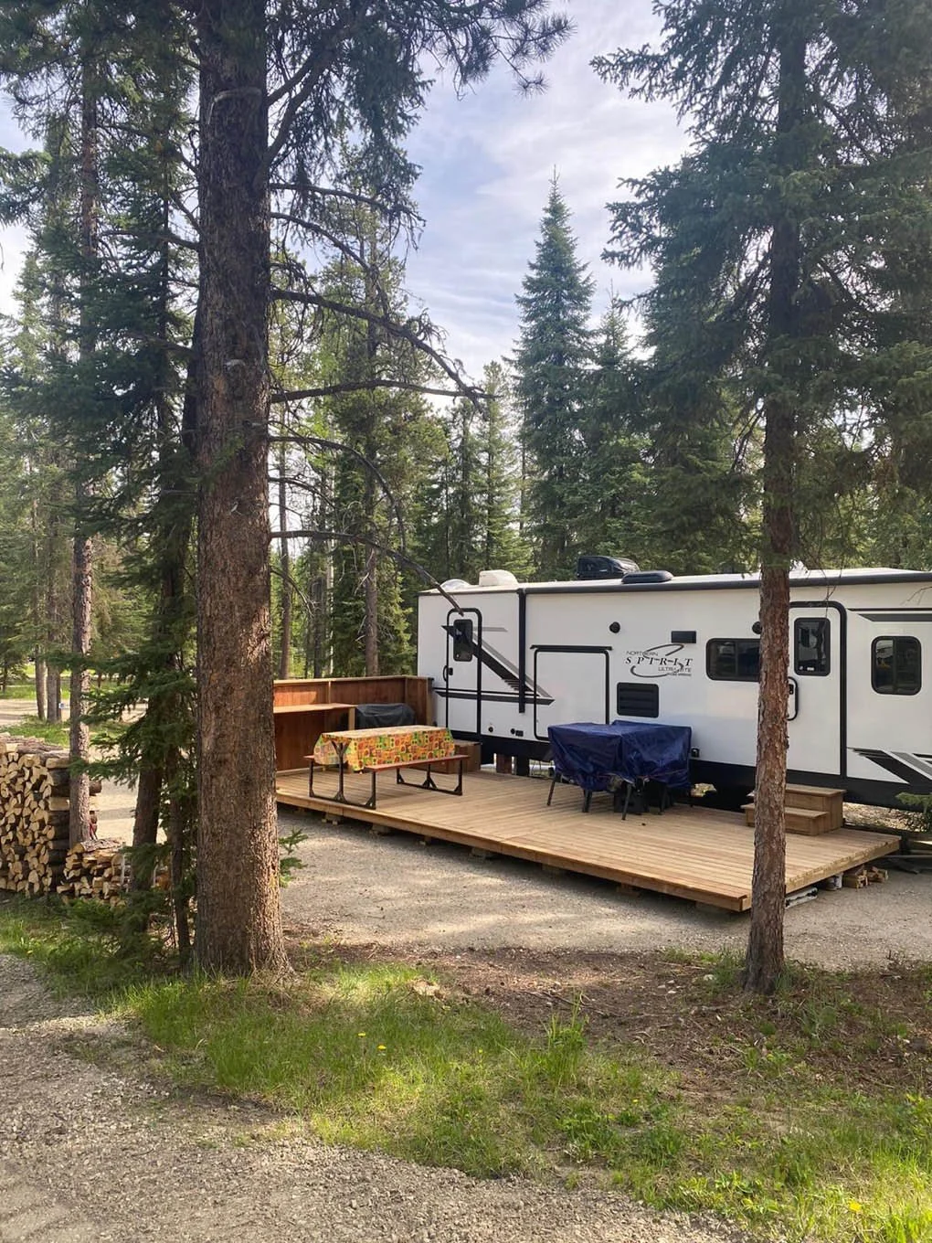 A travel trailer parked on a wooden deck in a forested area with trees and green foliage nearby.