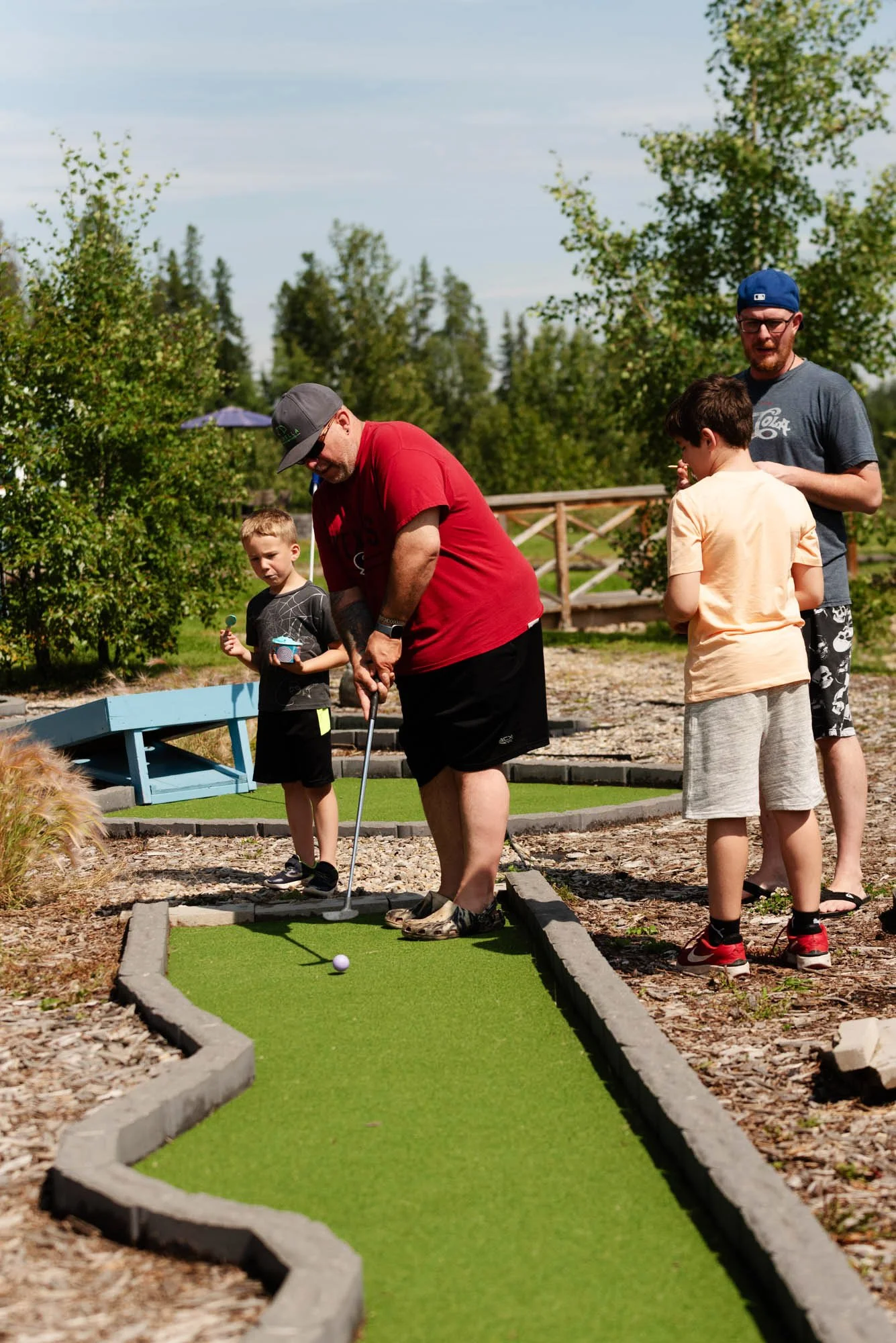 Four people playing mini golf outdoors, including a child holding a golf ball and a putter, a man aiming with a golf club, and two other boys watching, with trees and a wooden fence in the background.