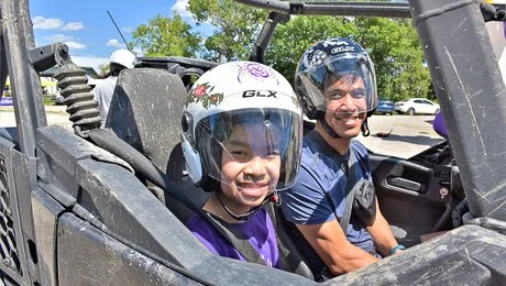 Two children wearing helmets sit in a side-by-side off-road vehicle with a black frame, outdoors on a sunny day.