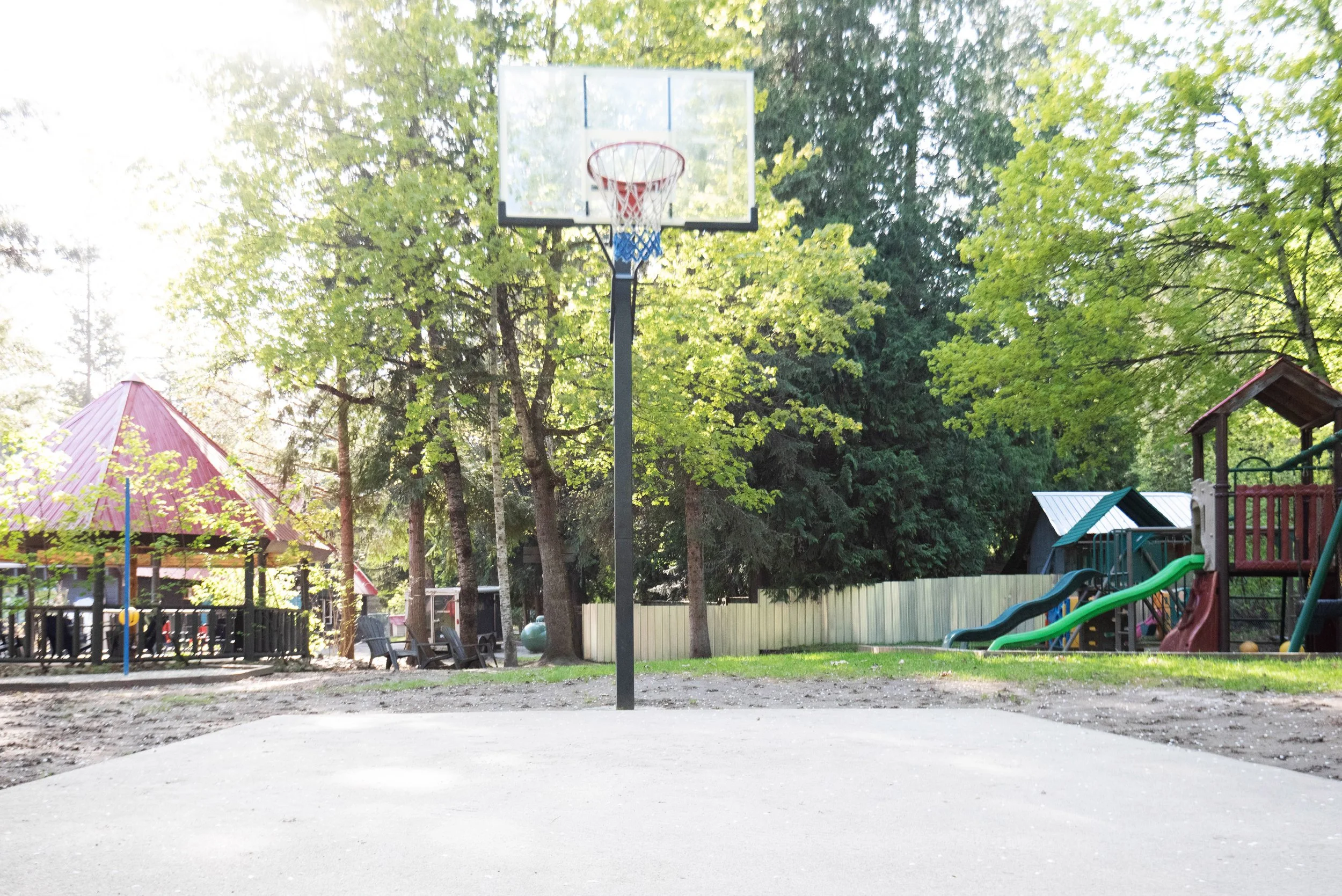 A playground with a basketball hoop in the foreground, surrounded by trees and playground structures, with a red roof and slides in the background.