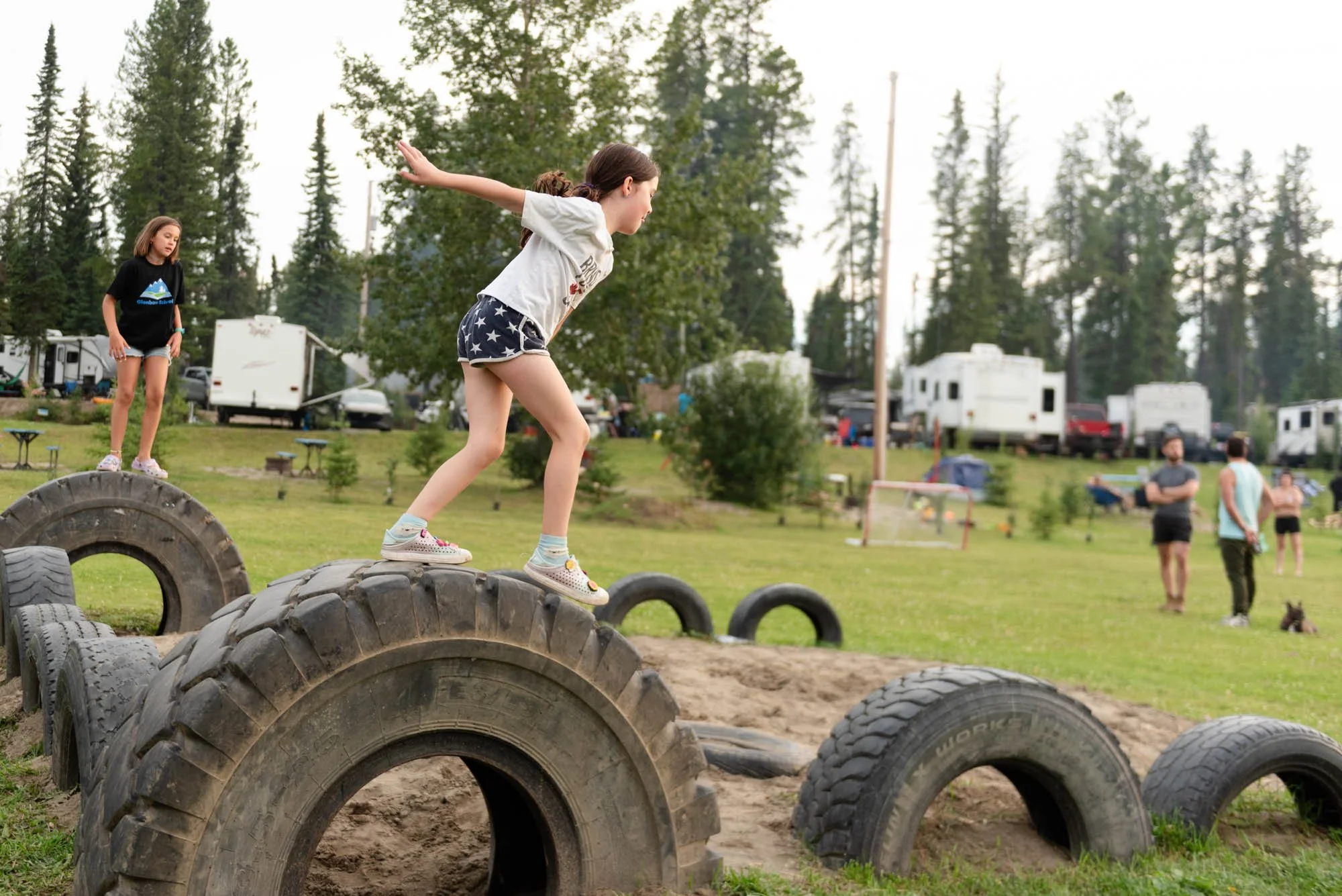 Two young girls are playing on large tires outdoors in a park or campground. One girl is balancing on the tires with arms outstretched, while the other girl stands on a tire watching her. In the background, there are trees, trailers, and a few adults