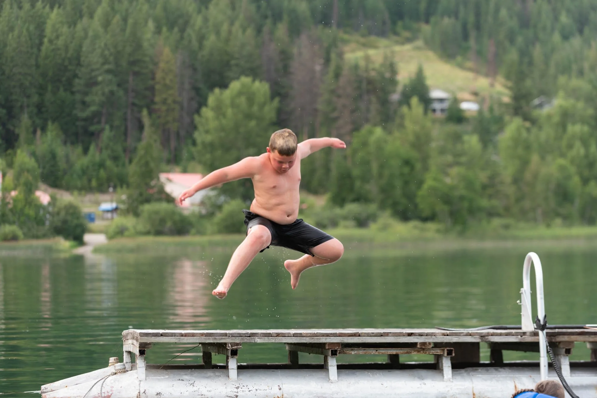 A shirtless boy in black swim trunks jumping off a dock into a lake with a forested hillside in the background.