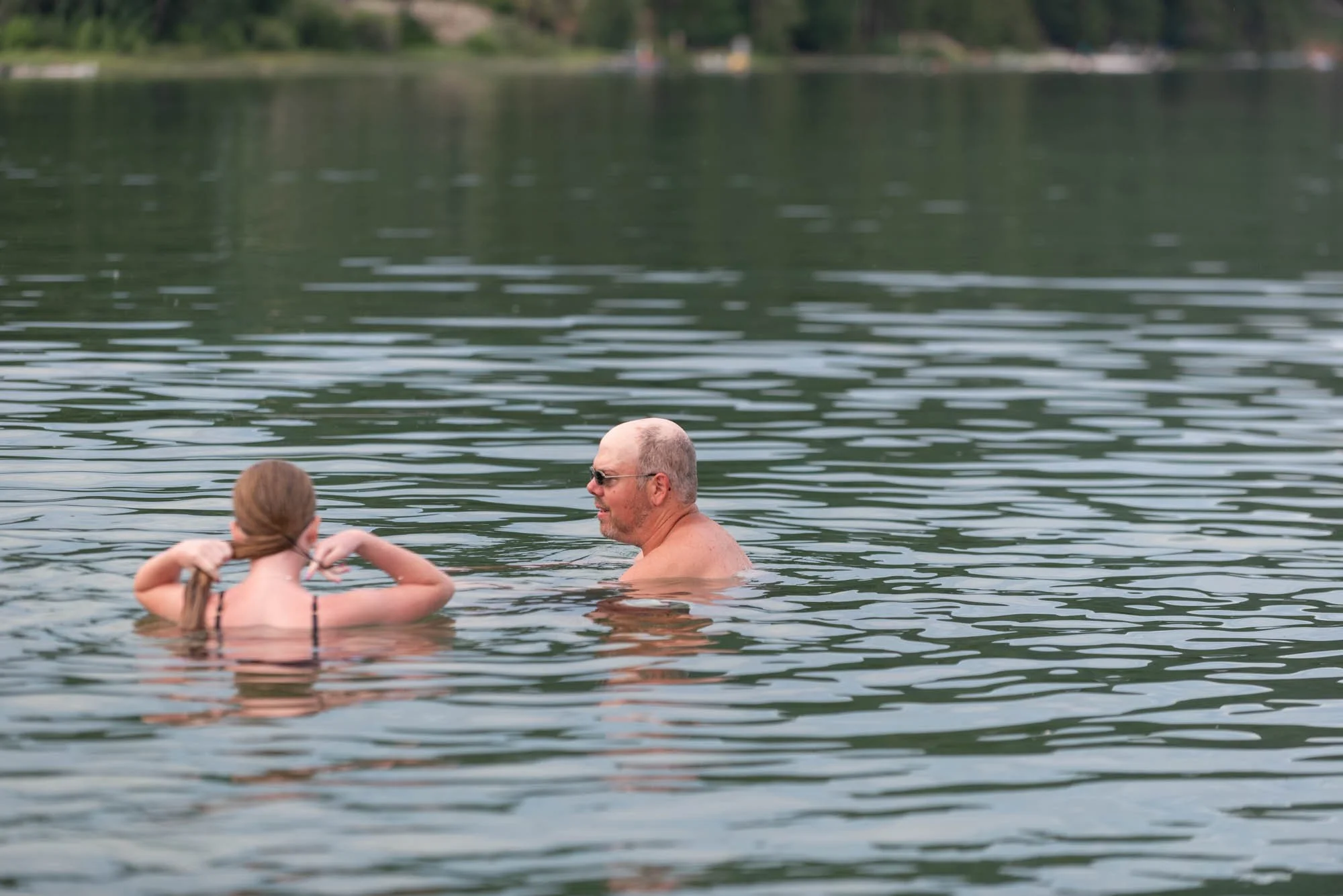 An older man with sunglasses swimming in a lake with a young girl, who has long hair, holding her hair back with her hands.