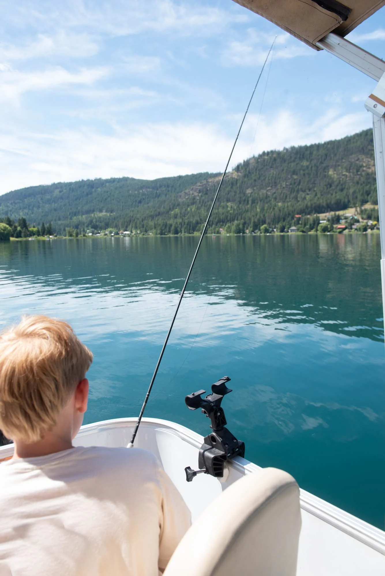 A young boy with blonde hair sitting in a boat, fishing in a calm lake with mountainous terrain and scattered houses in the background. The boat has a fishing rod and a mounted device.