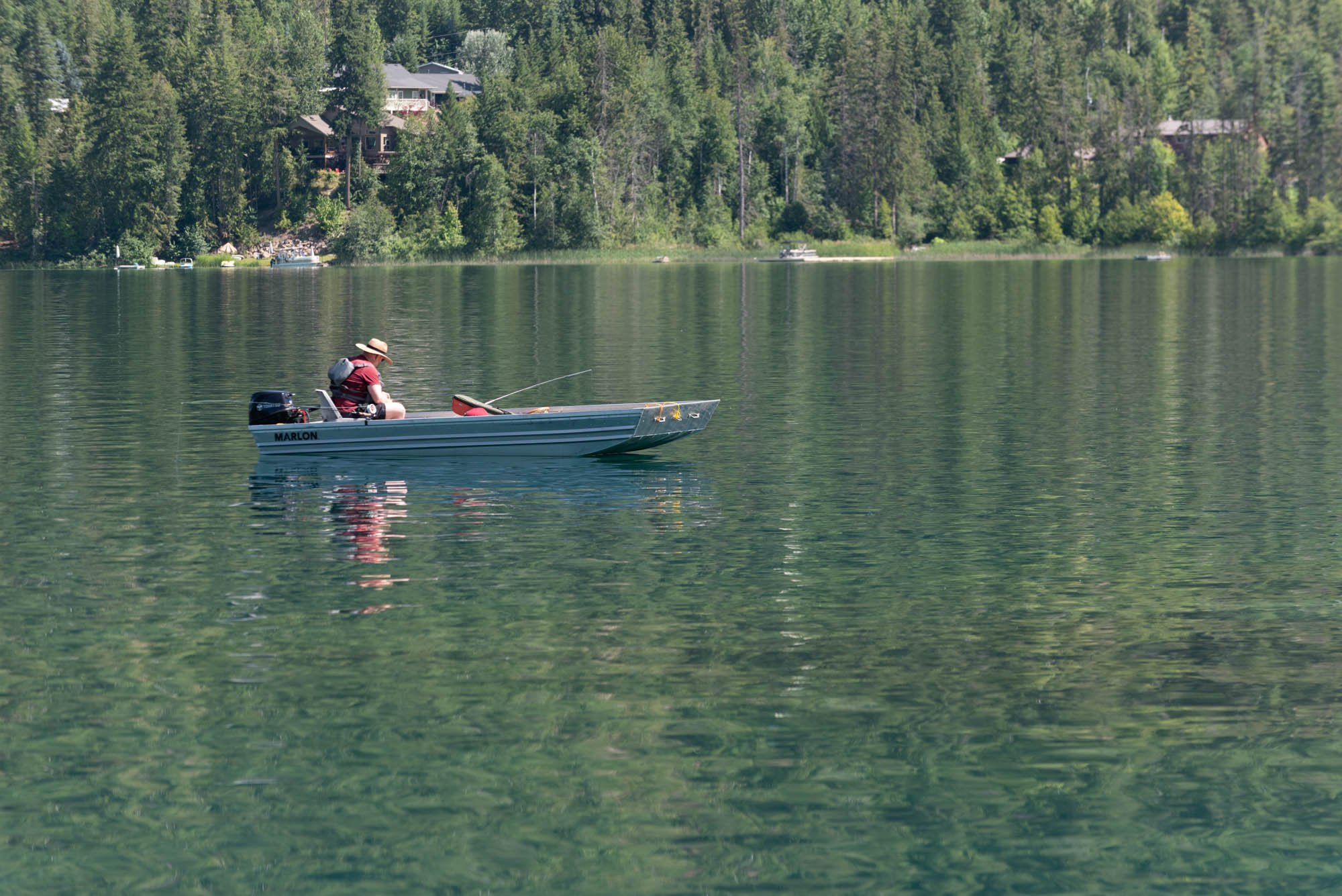 A person wearing a hat sitting in a small boat on a calm lake, surrounded by forested hills with houses visible on the hillside.