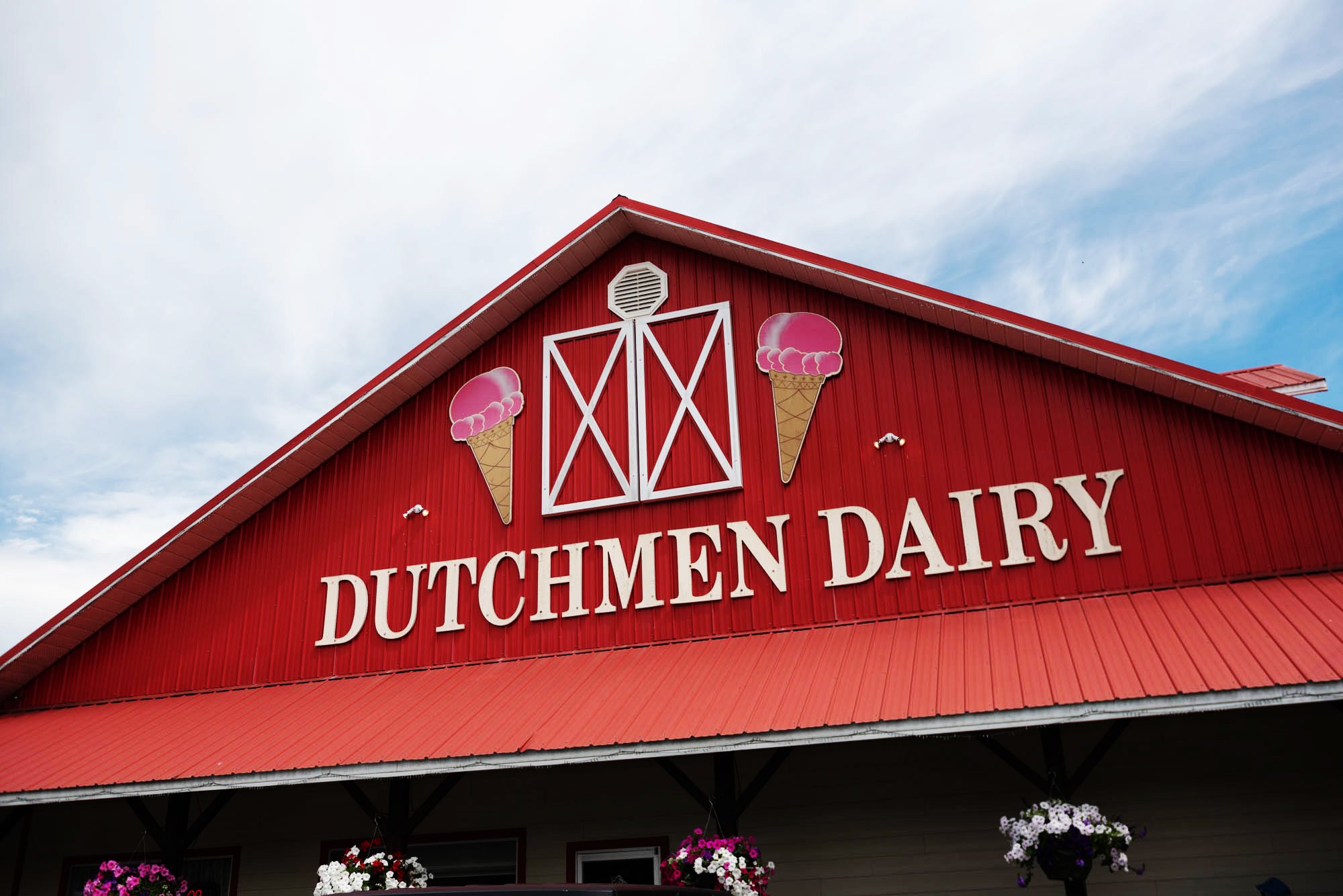 Red barn-style building with a sign that reads 'Dutchmen Dairy', decorated with ice cream cone images, and surrounded by hanging flower baskets.