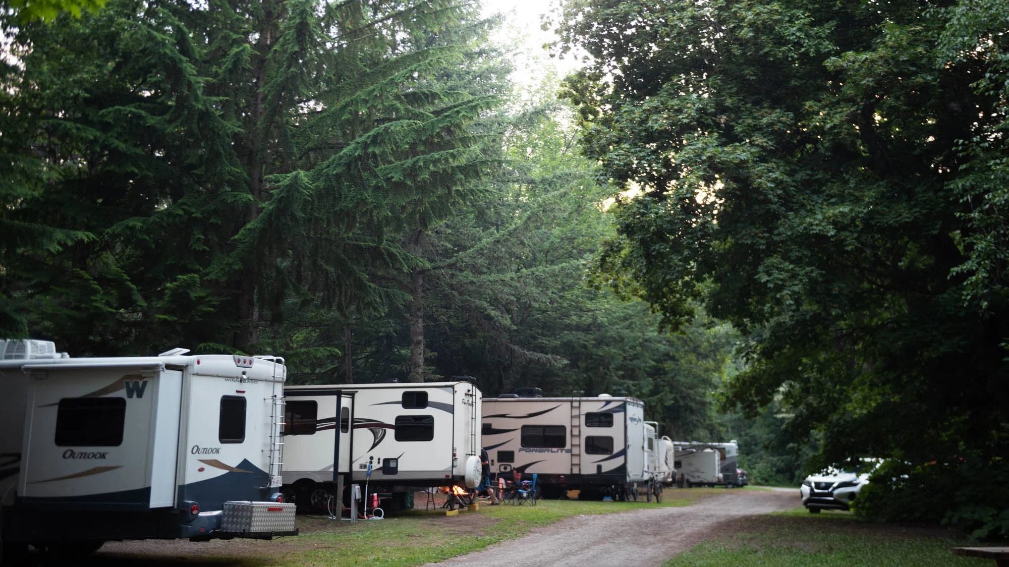 Camping area with multiple recreational vehicles (RVs) parked among tall green trees, with a gravel pathway and a partly cloudy sky.