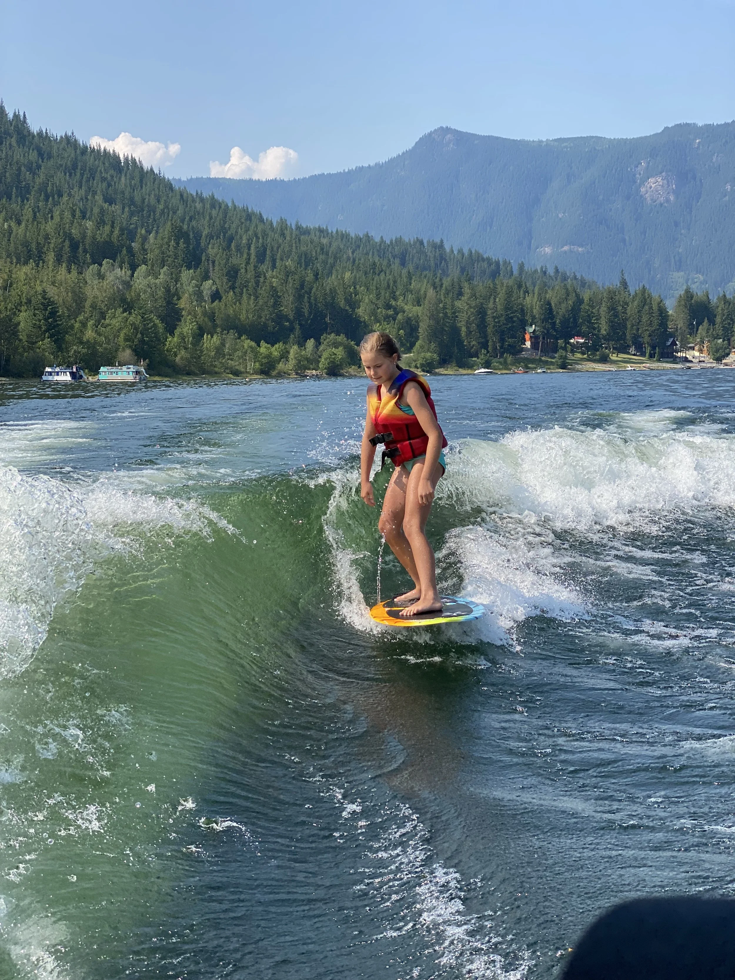 A girl in a red life jacket surfing on a wave in a lake surrounded by green wooded mountains.
