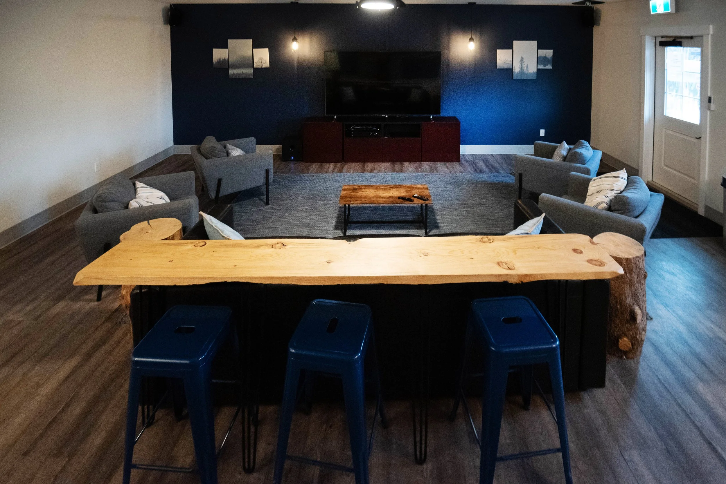 A cozy living room with a large flat-screen TV on a dark wood cabinet, surrounded by four gray armchairs and a wooden coffee table, with wood flooring and wall art.