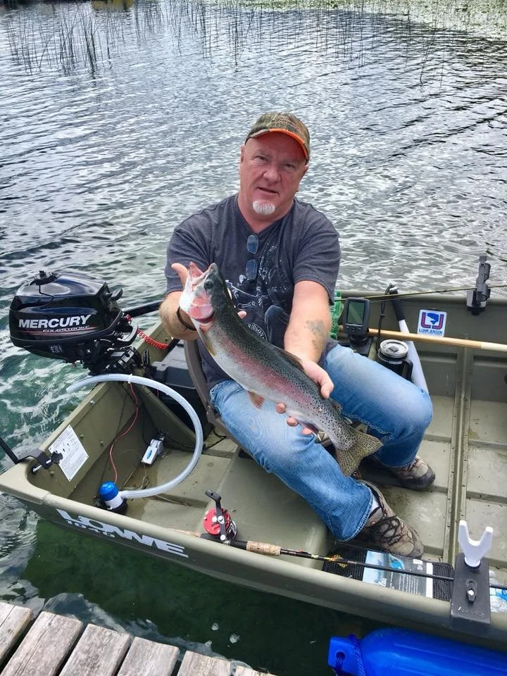 A man sitting in a small boat on the water holding a large rainbow trout he caught, with fishing gear and a Mercury outboard motor visible in the boat.