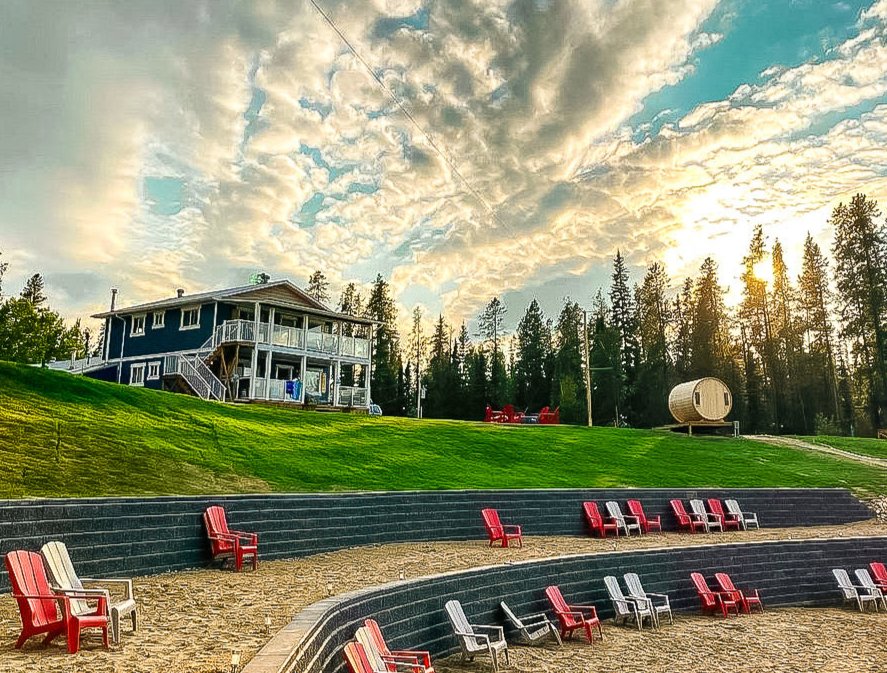 An outdoor amphitheater with red and white Adirondack chairs, a grassy hill, a two-story house, and a forest in the background during sunset.
