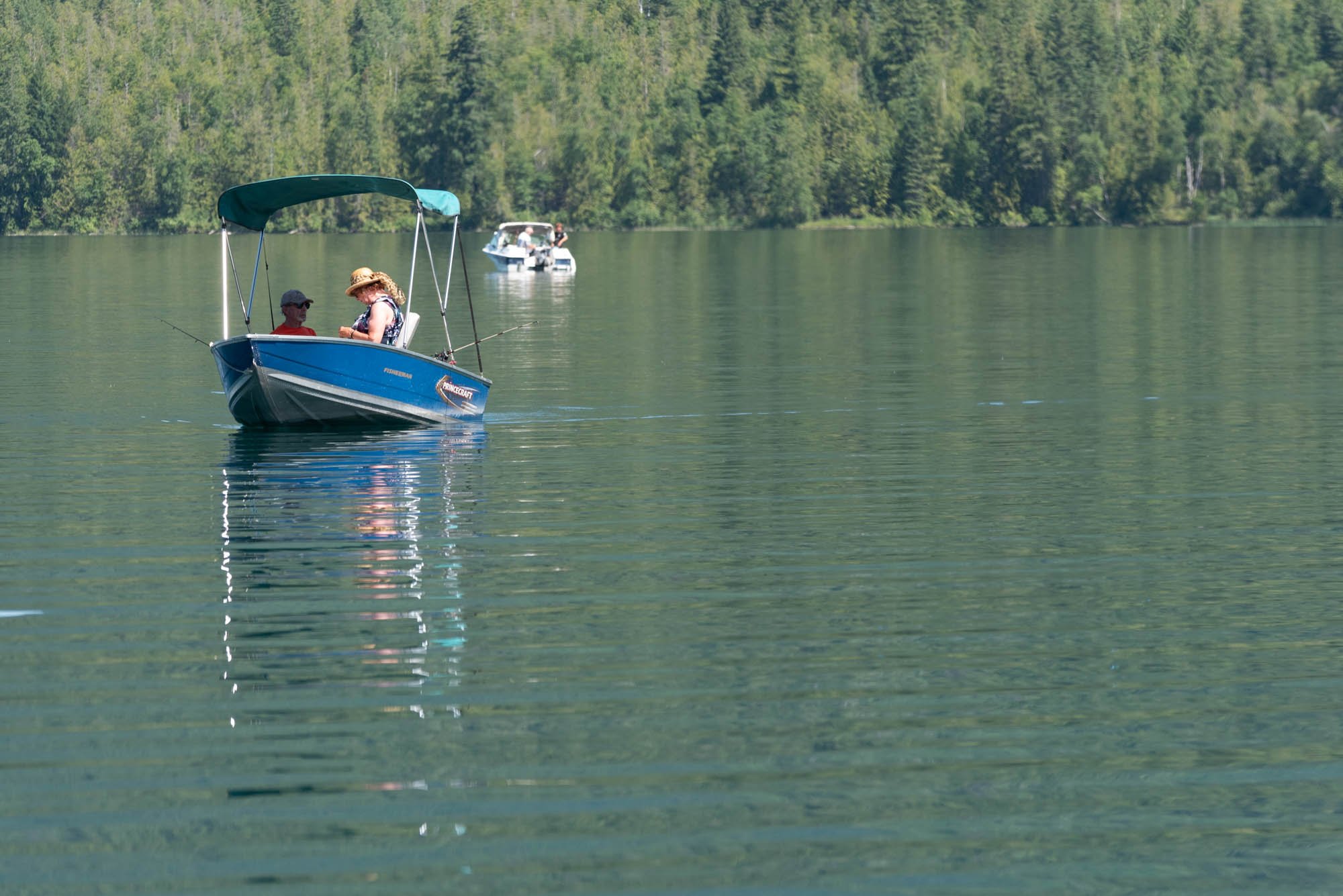 A small blue boat with a green canopy floating on calm lake water with three people fishing inside. Two more boats are visible in the background, and there is a lush green forest on the shoreline.