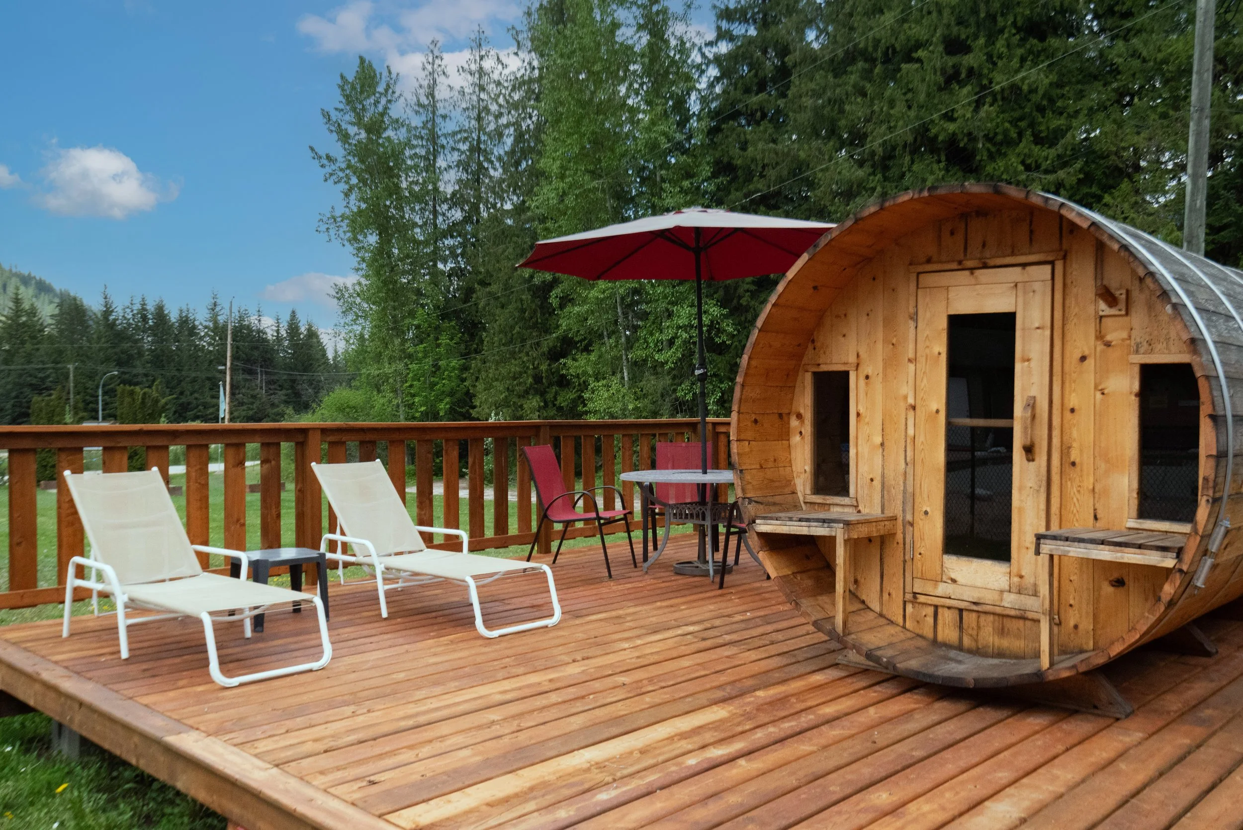 A wooden deck with lounge chairs, a table with an umbrella, and a small wooden curved cabin, set outdoors with green trees and a partly cloudy sky in the background.