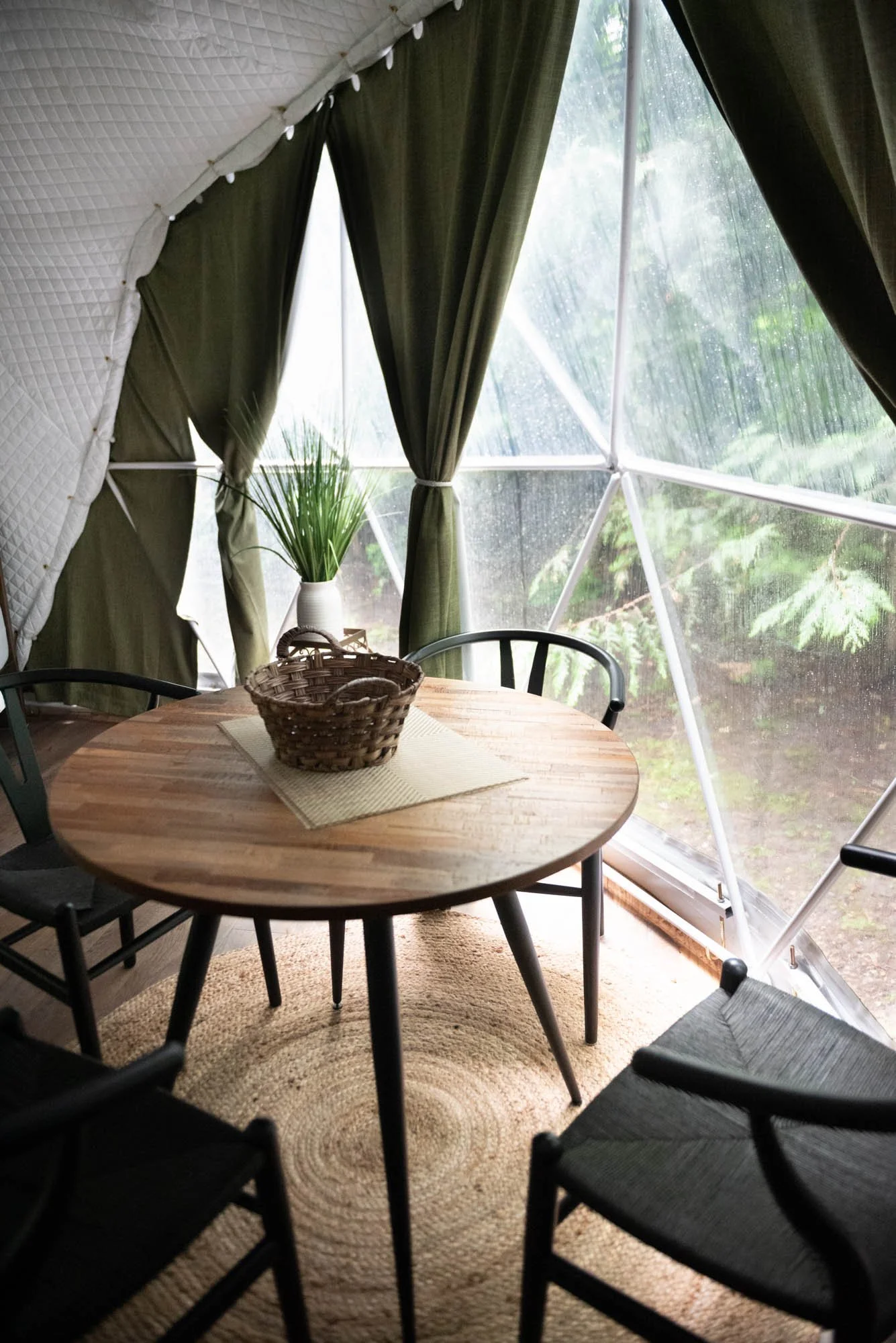 A round wooden dining table with a woven basket and placemat, surrounded by black chairs, inside a tent with green curtains and a rain-spattered window showing greenery outside.