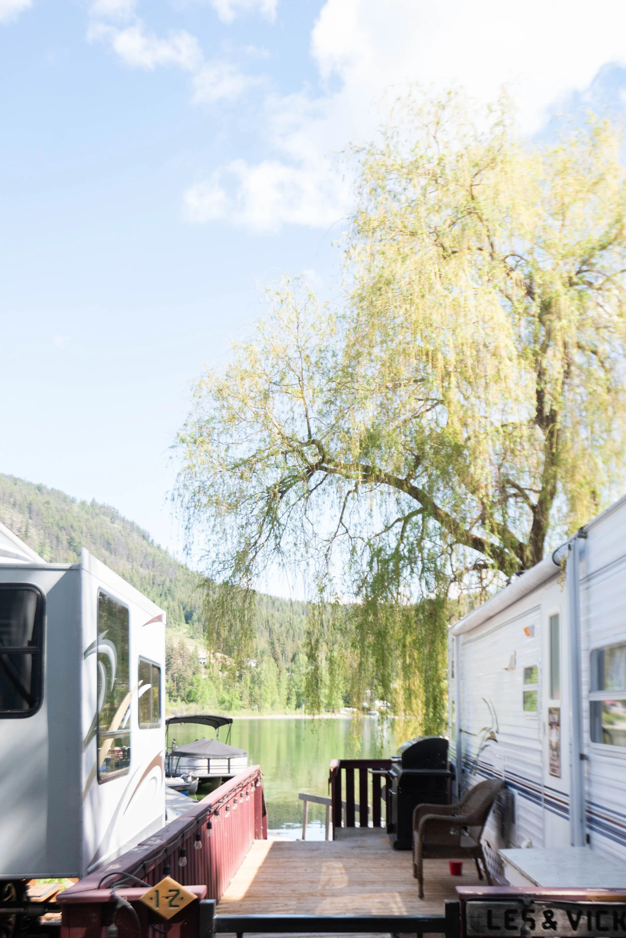 View of a lakeside campsite with RVs, outdoor furniture, a boat, and a large tree under a partly cloudy sky.