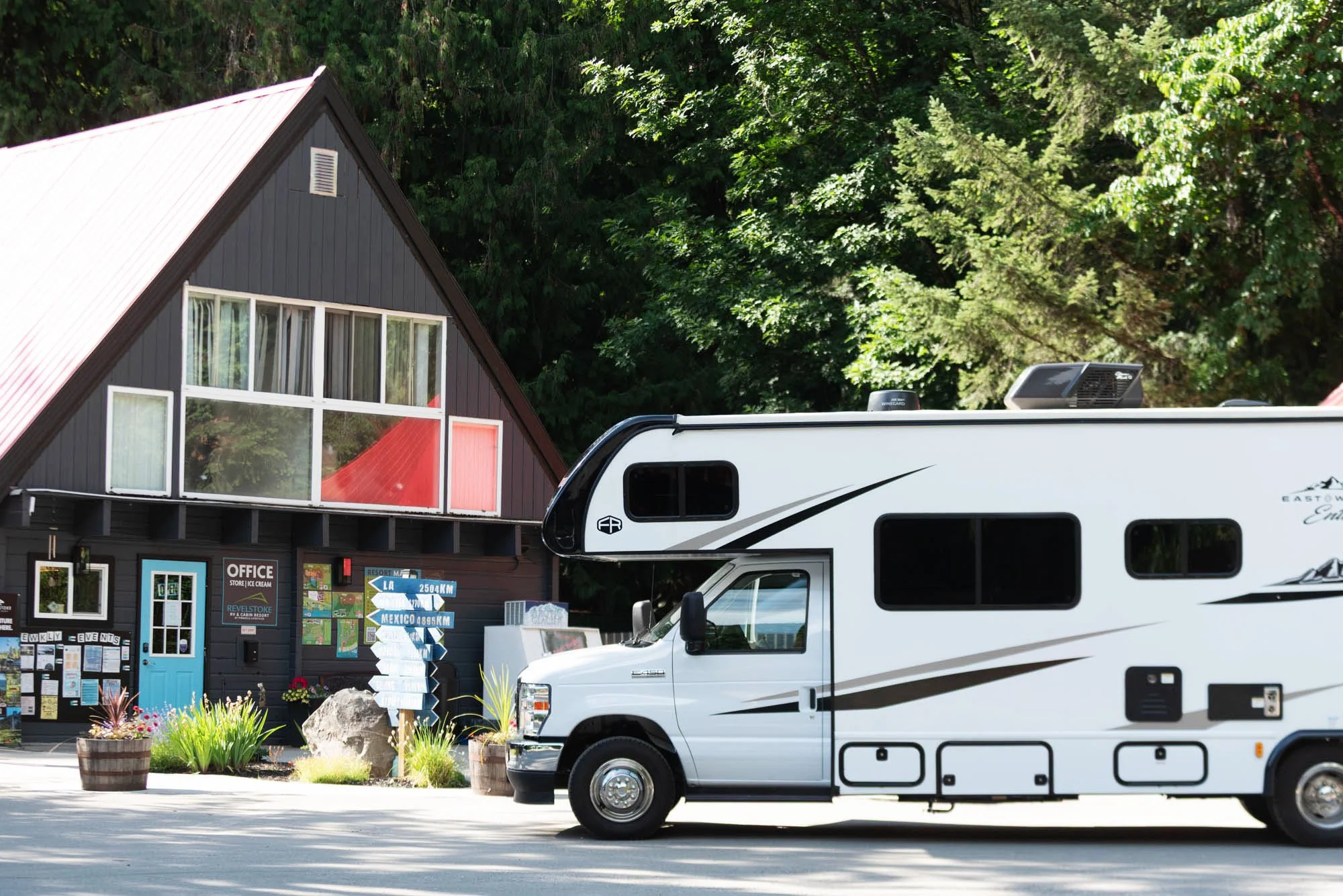 A black A-frame building with large windows, outdoor signs, and plants in pots, alongside a white motorhome parked in front, with a forest of tall green trees in the background.